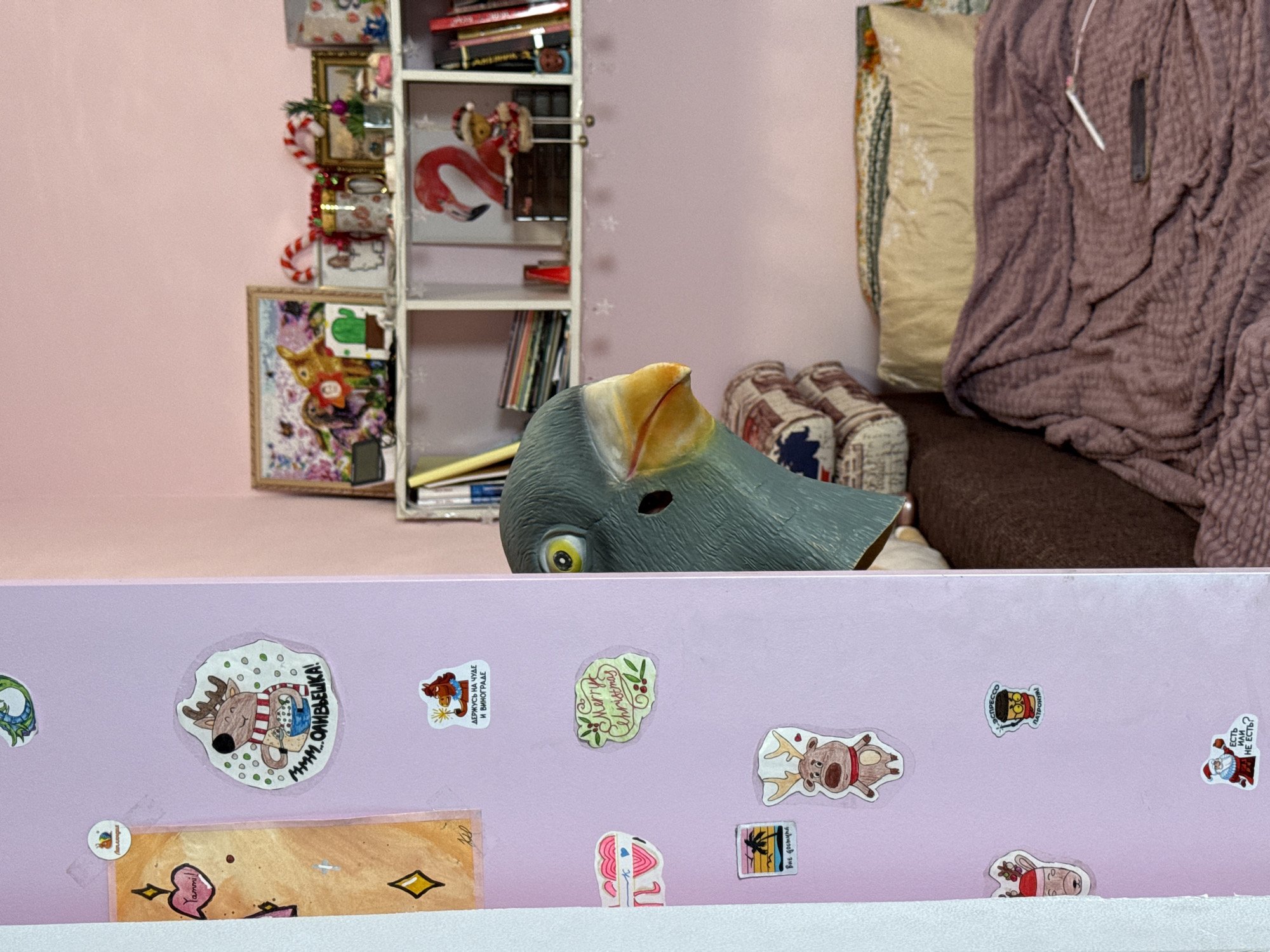 Here is a forensic description of the image:
The image presents a close-up scene with several distinct layers. In the foreground, a pink surface is prominently displayed, decorated with various stickers featuring cartoonish characters and holiday-themed designs. Some of the stickers have Cyrillic text, adding cultural context.
Behind this pink surface is a pale gray rubber or plastic object that appears to be a partial animal mask, possibly of a bird. It has a textured surface and a yellow-orange beak.
In the background, a miniature shelving unit stands, adorned with small decorative items, including framed pictures and what appear to be miniature books. To the right, a cushioned area with pillows and a draped blanket in earth tones provides additional depth to the scene.
The scene's location cannot be definitively determined to be in Rostov-on-Don, Russia, without additional information or visual cues confirming the location.