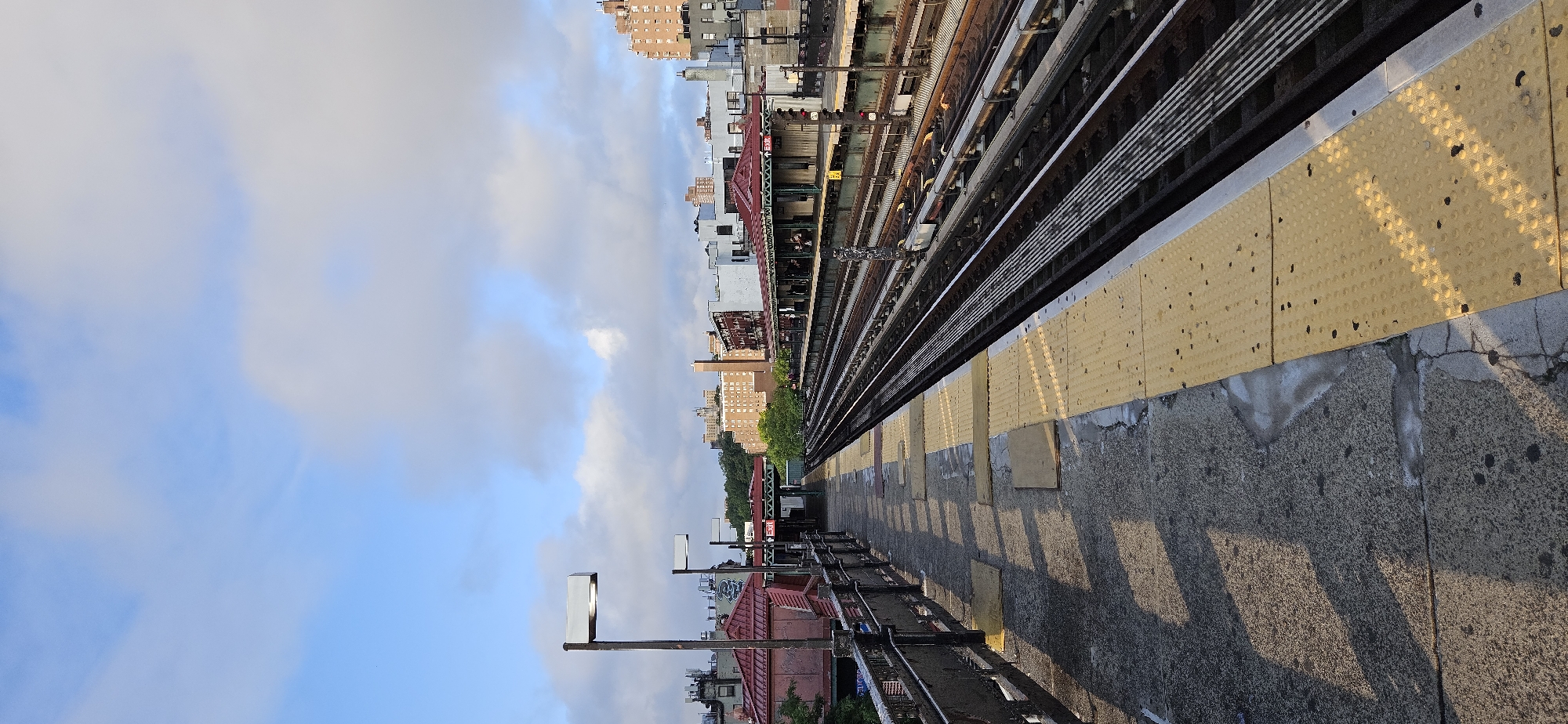 This is a wide-angle, eye-level shot of an elevated subway platform in Queens, New York City, on a partly cloudy day. The platform, made of concrete with yellow tactile warning strips along the edge, runs diagonally from the lower left to the upper right of the frame. The tracks of the elevated subway line run parallel to the platform on the right. In the background, a line of buildings, including apartment complexes and some red-roofed structures, are visible against a pale blue sky with scattered white clouds. There are no people clearly visible in the image.