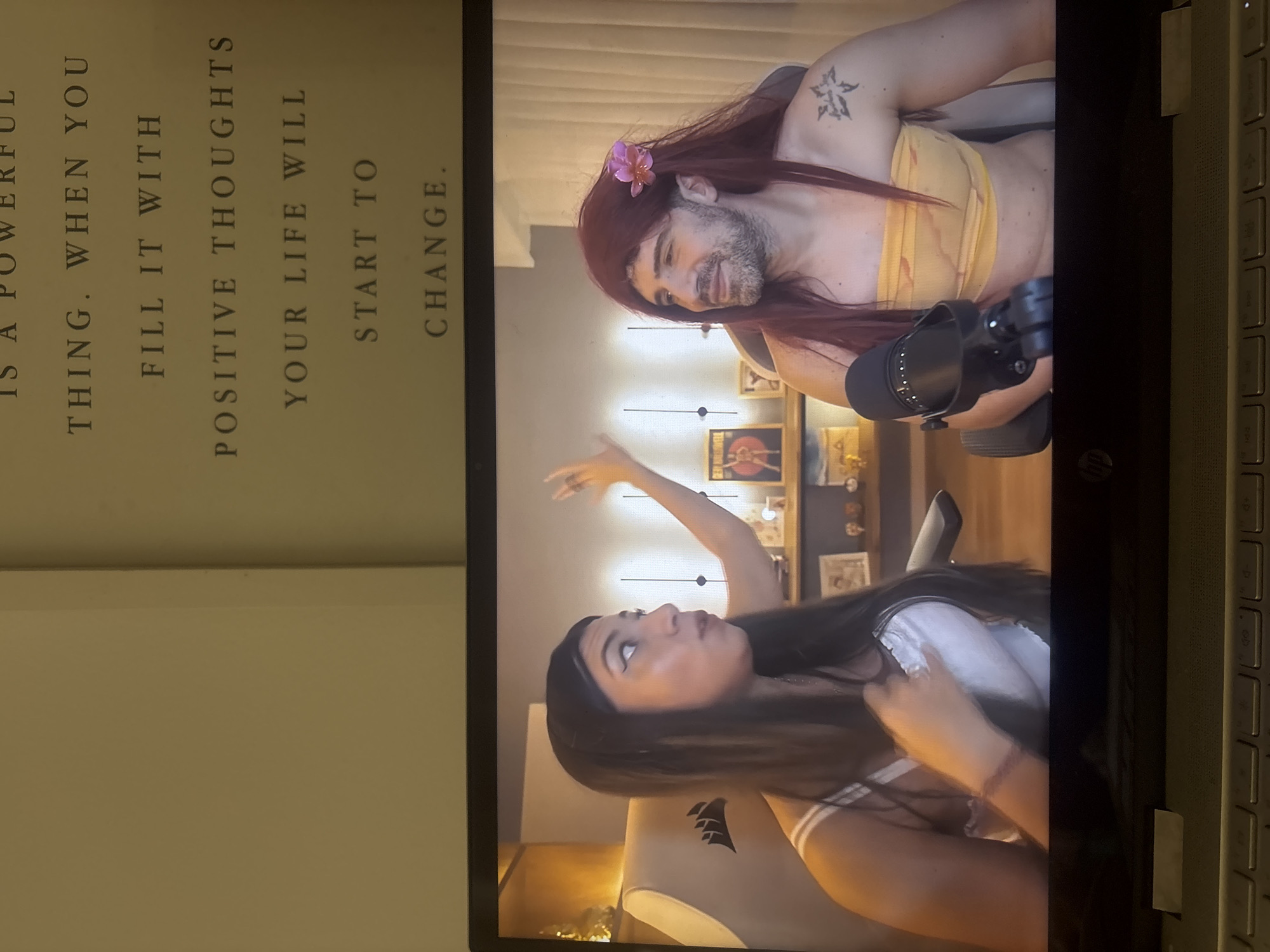 An image captures a laptop screen displaying two individuals engaged in what appears to be a video call or livestream, positioned next to an open book or paper with text.
On the laptop screen, a man is visible on the right side, sporting a beard and long reddish-brown hair adorned with a pink flower. He is wearing a yellow tube top and has a tattoo on his left shoulder. He looks downward with a subtle smile. A microphone stands in front of him. On the left side of the screen, a woman with long dark hair is seen looking upwards and gesturing with her right hand. She is wearing a white top, possibly a school uniform style. Both appear to be seated in chairs, with a "Corsair" logo visible on the woman's chair. The background behind them is an indoor setting, softly lit, with framed pictures on the wall.
Adjacent to the laptop, a page is visible, displaying the text: "IS A POWERFUL THING. WHEN YOU FILL IT WITH POSITIVE THOUGHTS YOUR LIFE WILL START TO CHANGE." The laptop itself bears an "hp" logo. The man's attire and wig suggest a playful or costume-themed interaction. The setting suggests an indoor environment, possibly a home studio.