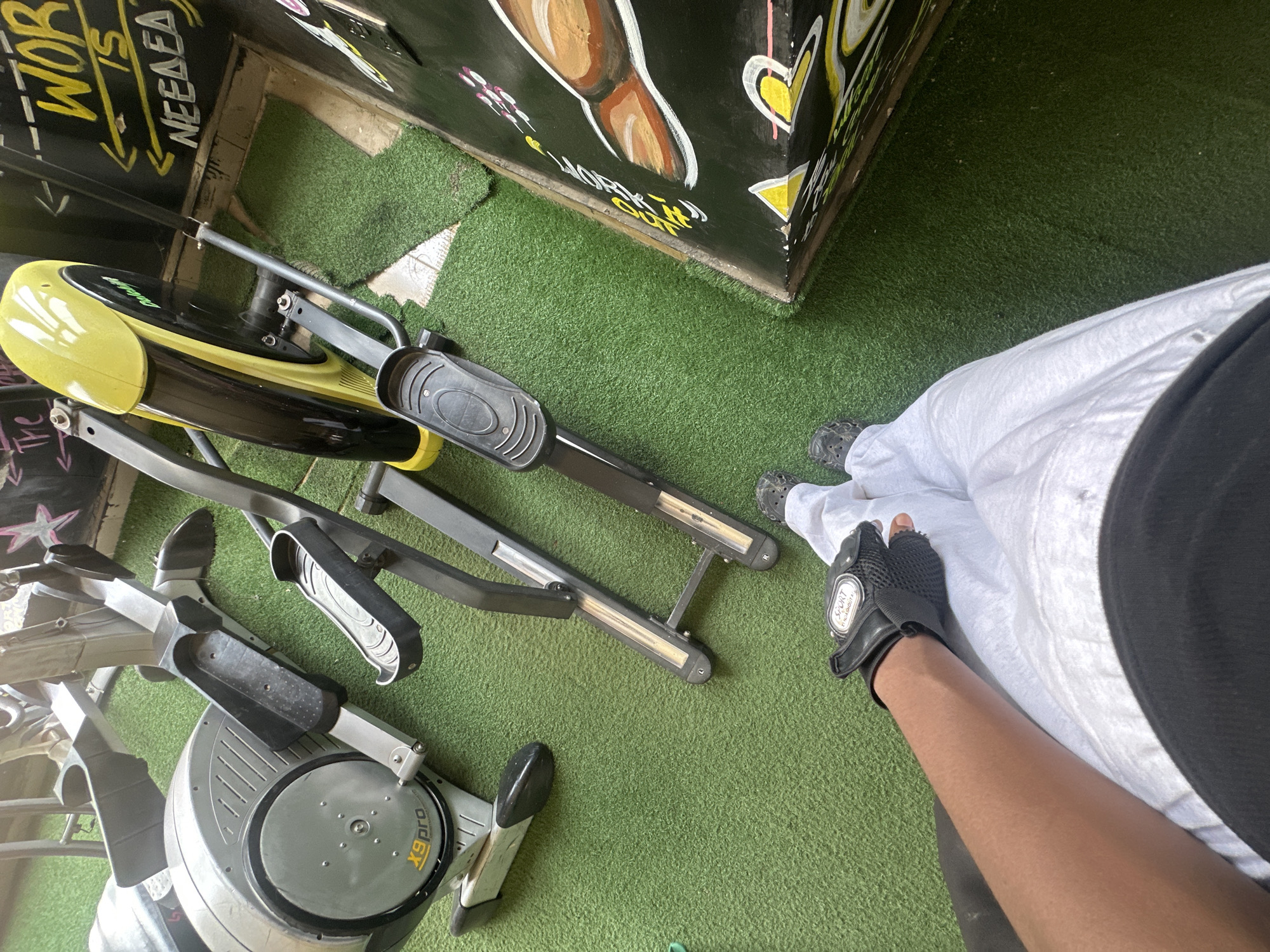 Scene: Indoor space, likely a gym in Iddo Tudunwada, Nigeria.
Objects: Two elliptical machines (one yellow/black, one gray), artificial green turf flooring, painted wall with graffiti-style writing and drawings, a person wearing a white garment, black top, and a black fingerless glove.
People: One person partially visible.
Actions: The person is standing near the elliptical machines.
Environment: The scene suggests a workout area or a small gym.