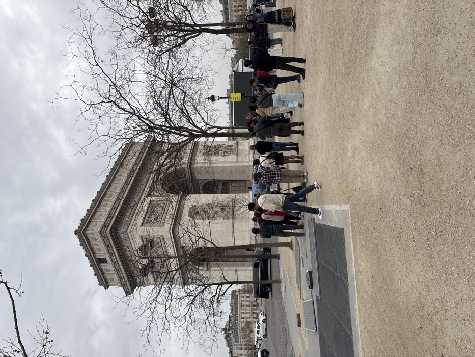 The scene depicts a daytime view of the Arc de Triomphe, located at Place Charles de Gaulle in Paris, France, under an overcast sky. The foreground consists of a light brown, unpaved ground surface, which transitions to a gray paved area on the left featuring several dark, rectangular metal grates. Bare deciduous trees with slender branches are scattered throughout the midground, partially obscuring the lower sections of the monument.
Approximately 20-25 individuals, primarily adults, are gathered in the midground, standing on the unpaved surface. Most are facing generally towards the Arc de Triomphe, appearing to observe the monument. One male individual, wearing a light-colored hooded sweatshirt, blue jeans, and white athletic shoes with black stripes, is walking from left to right in the mid-left foreground. To the left of the monument, a paved street is visible with a black minivan and at least two white passenger cars. Distant Parisian buildings are discernible on the far left horizon. A black street lamp with a rectangular yellow sign is visible among the trees to the right of the gathered people.