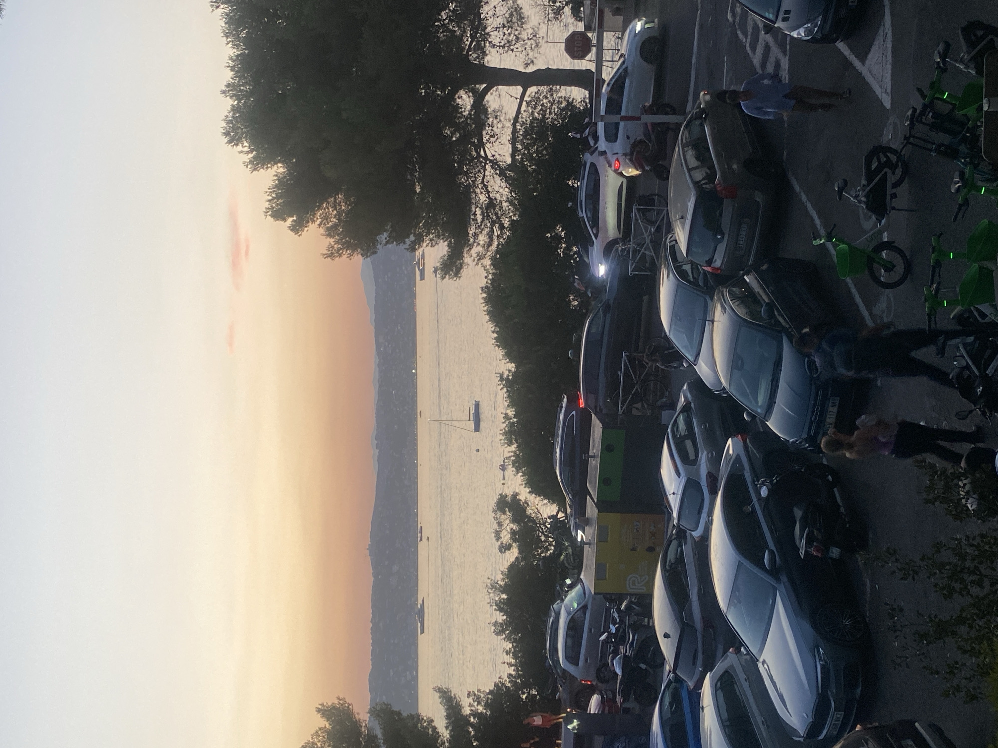 A high-angle, full shot shows a parking lot filled with numerous cars and motorcycles at dusk, overlooking a body of water with a distant coastline. The sky is a gradient of pale yellow to light orange, with a few pink clouds visible. A large pine tree dominates the right side of the frame.
In the foreground, a dark gray BMW sedan is parked, with a black scooter parked behind its rear bumper. To the right of the BMW, a woman in a pink tank top and dark pants walks away from the camera, carrying a baby. Further right, a man in a blue shirt and brown pants walks away from the camera. In the middle of the parking lot, a light green electric bicycle with a basket is parked. Several other cars and motorcycles are visible throughout the lot.
In the middle ground, various cars are parked. A silver SUV is parked to the right of a dark gray hatchback. A white SUV is parked to the right of the silver SUV, with its headlights on. Further back, a dark gray hatchback is parked to the left of a silver hatchback. A dark gray sedan is parked to the left of the silver hatchback.
The background shows a body of water, likely the Mediterranean Sea, with several boats and yachts on it. A sailboat is visible in the water to the right of a larger yacht. The distant coastline is lined with hills and buildings, appearing hazy in the dusk light. A small hill with a structure on top is visible on the far right of the distant coastline. The image appears to have been taken in Antibes, France, judging by the style of the cars and the scenery.