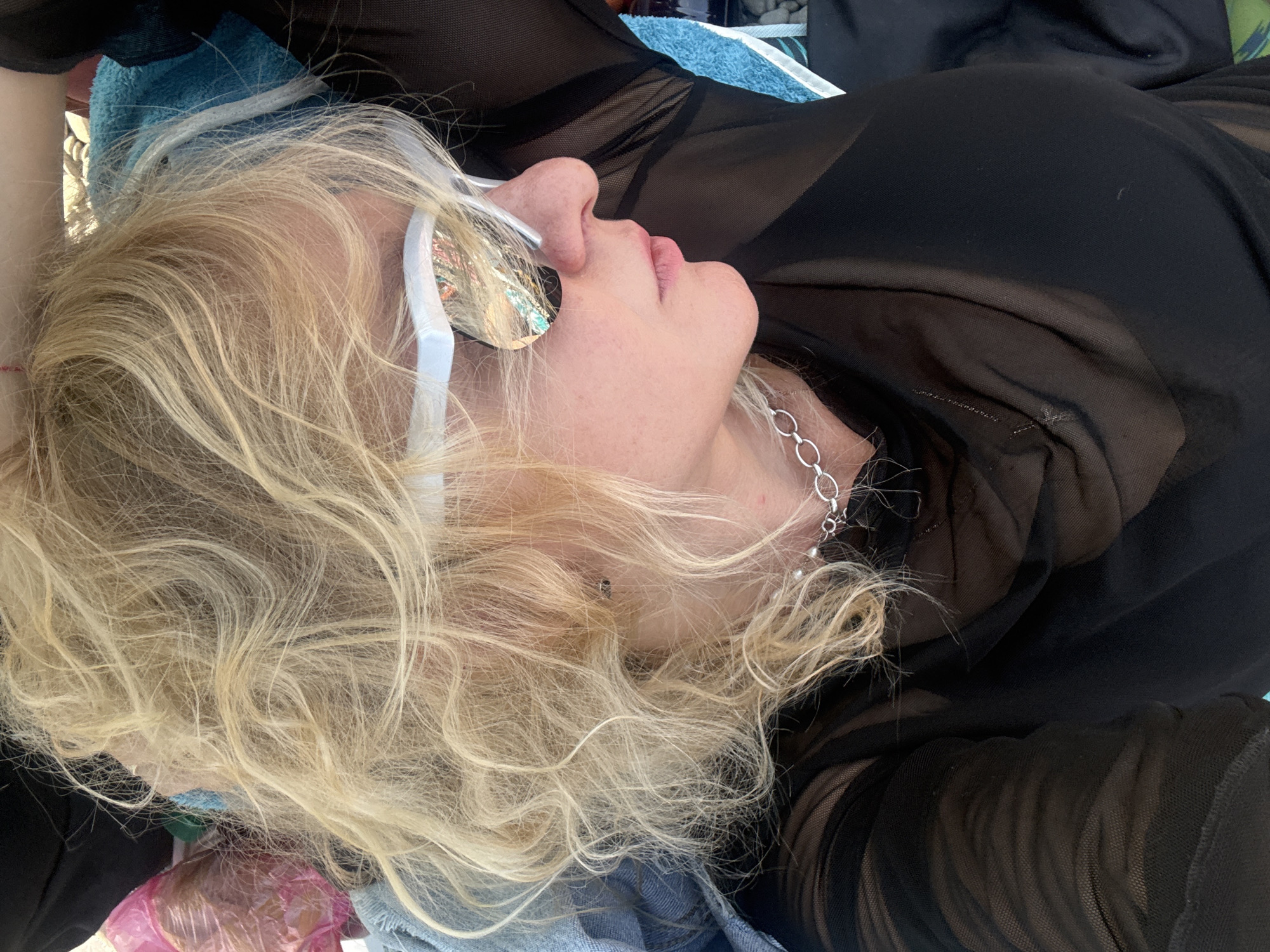 A woman with blonde, curly hair, wearing white-rimmed sunglasses and a sheer black top, lies on a blue towel, possibly at a beach or pool area in Partenit, Ukraine. Her head is tilted upwards, and she is looking to her right. Her right arm is bent upwards, resting behind her head. A silver chain necklace is visible around her neck. The sunglasses reflect the surrounding environment, which appears to be sunny and outdoor.