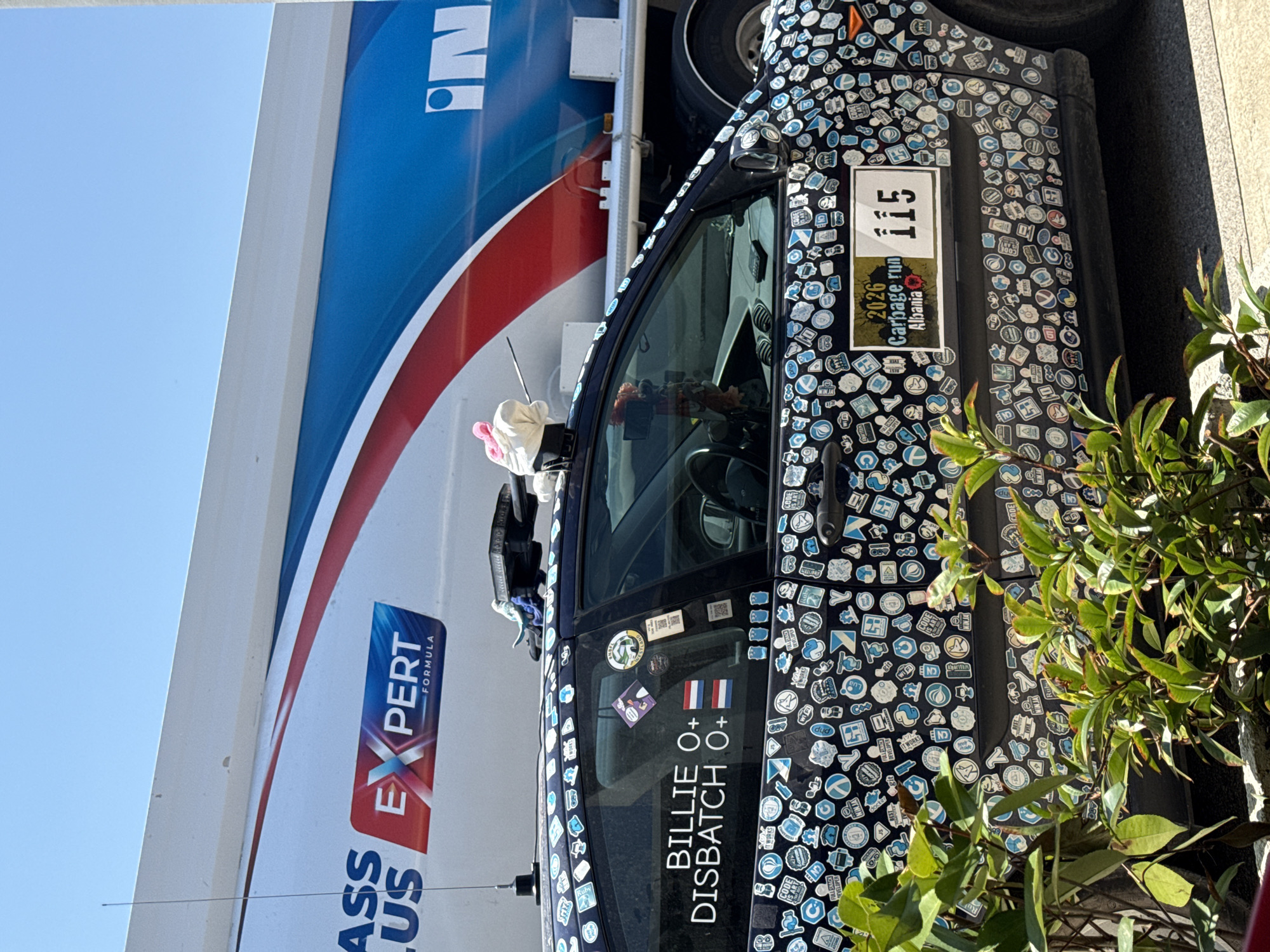 This image shows the side of a dark-colored car heavily adorned with numerous small stickers, many of which appear to be related to technology and programming. A larger sticker on the driver's side door displays "115" and "2026 Garbage run Albania," suggesting the car might be participating in a rally or event. Adjacent to this, text reads "BILLIE O+ DISBATCH O+" with two small flag stickers, one of which is the Dutch flag.
In the background, a large white truck with blue and red stripes is partially visible. The truck has "IN" and "EXPERT FORMULA" text on its side. The scene appears to be outdoors during the daytime, with clear blue sky visible on the left. Some green foliage is present in the foreground on the right, partially obscuring the car and truck. The overall impression is of a custom-decorated vehicle, possibly at a stop during a road trip or event. No people are clearly visible in the frame. The lighting suggests it is a sunny day.
