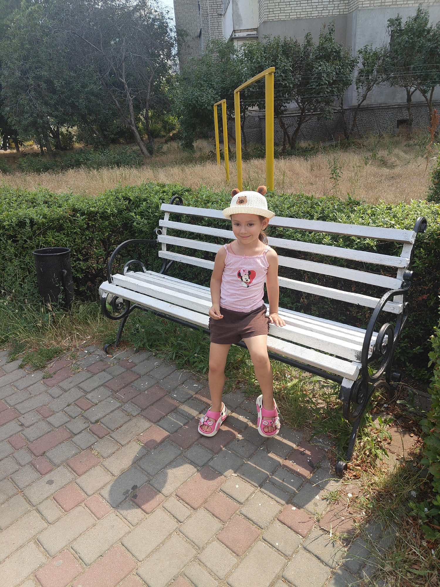 A young girl sits on a white park bench, facing forward with a slight smile. She wears a white straw hat with bear ears, a pink tank top with a Minnie Mouse graphic, and brown shorts. Her pink sandals have white flower details. The bench has a black metal frame and is set against a backdrop of dense green bushes. To the left of the bench, a black trash can stands on a paved walkway made of red and gray bricks. Beyond the bushes, tall dry grass and trees are visible, with a portion of a grey brick building in the upper right corner. Yellow poles, possibly part of a playground, are also present in the background. The scene suggests a sunny day, likely in the late morning or early afternoon, given the shadows. The girl's expression and posture indicate a relaxed and content mood. There is no visible text in the image.