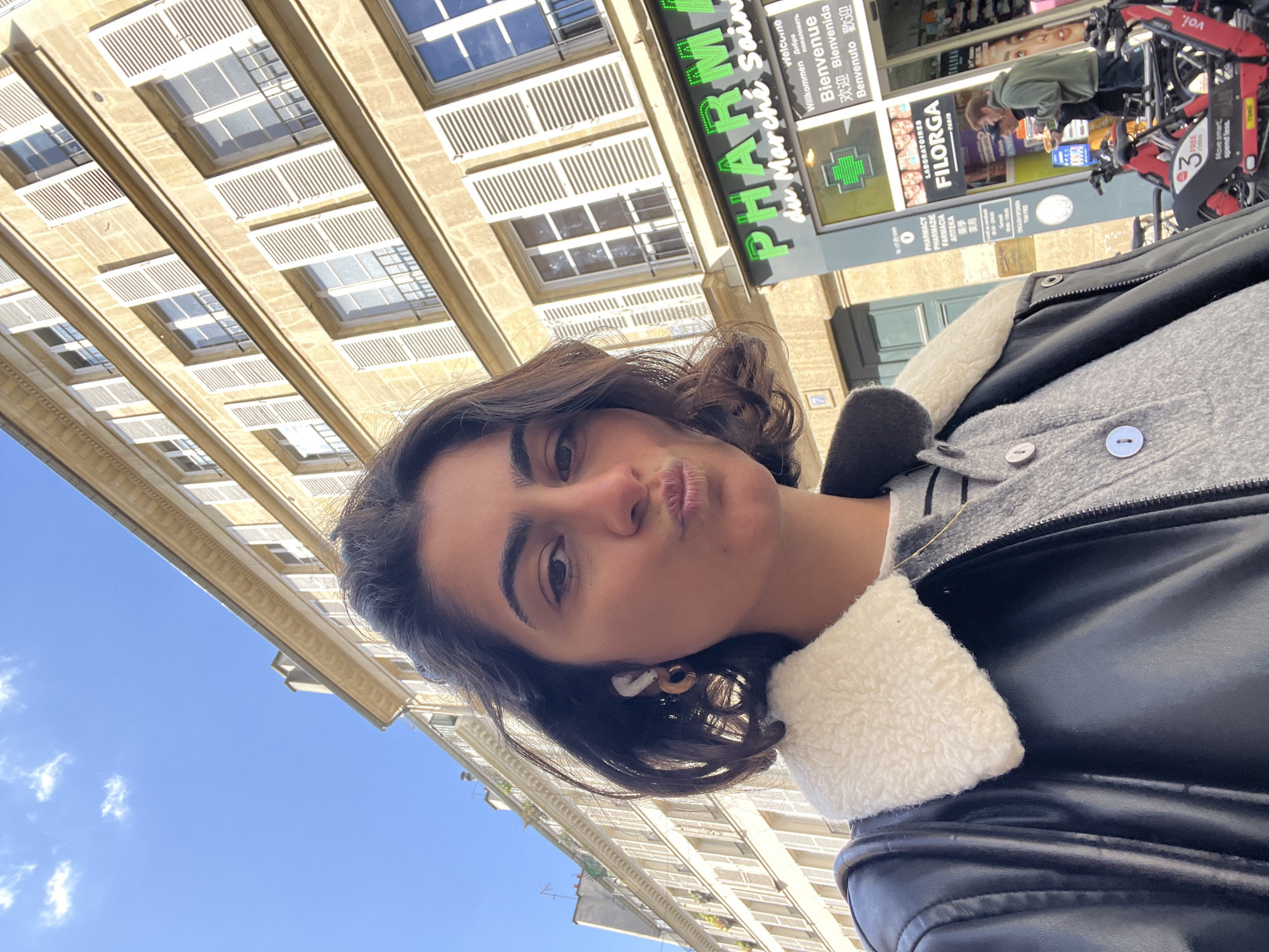 A selfie taken by a young woman with dark hair and brown eyes. She is wearing a gray jacket with white fur trim under a black leather jacket. She is making a kissing face. The photo appears to be taken in Paris, France, as evidenced by the "PHARMA du Marché Saint" sign in the background and French text. There is a bike rental station, possibly "voi," in the background as well. The architectural style suggests a European urban setting. The weather is sunny with a blue sky visible.