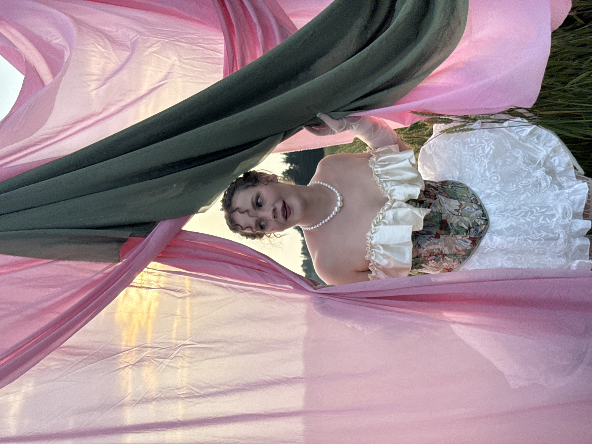 In Malyi Bukor, Russia, a young woman is pictured in an outdoor setting, seemingly a field of grain, during what appears to be sunset. She is partially obscured by flowing fabrics, one pink and one dark green, draped and held to frame her. The woman is wearing a white pearl necklace, an off-the-shoulder ivory top with ruffled detailing, a patterned corset, and a textured white skirt. She is also wearing sheer gloves. Her gaze is directed to the left, and her mouth is slightly open as if speaking or reacting. The background reveals a hazy sky with warm light, indicative of the golden hour, and a line of trees in the distance. The overall impression is a theatrical or staged scene.