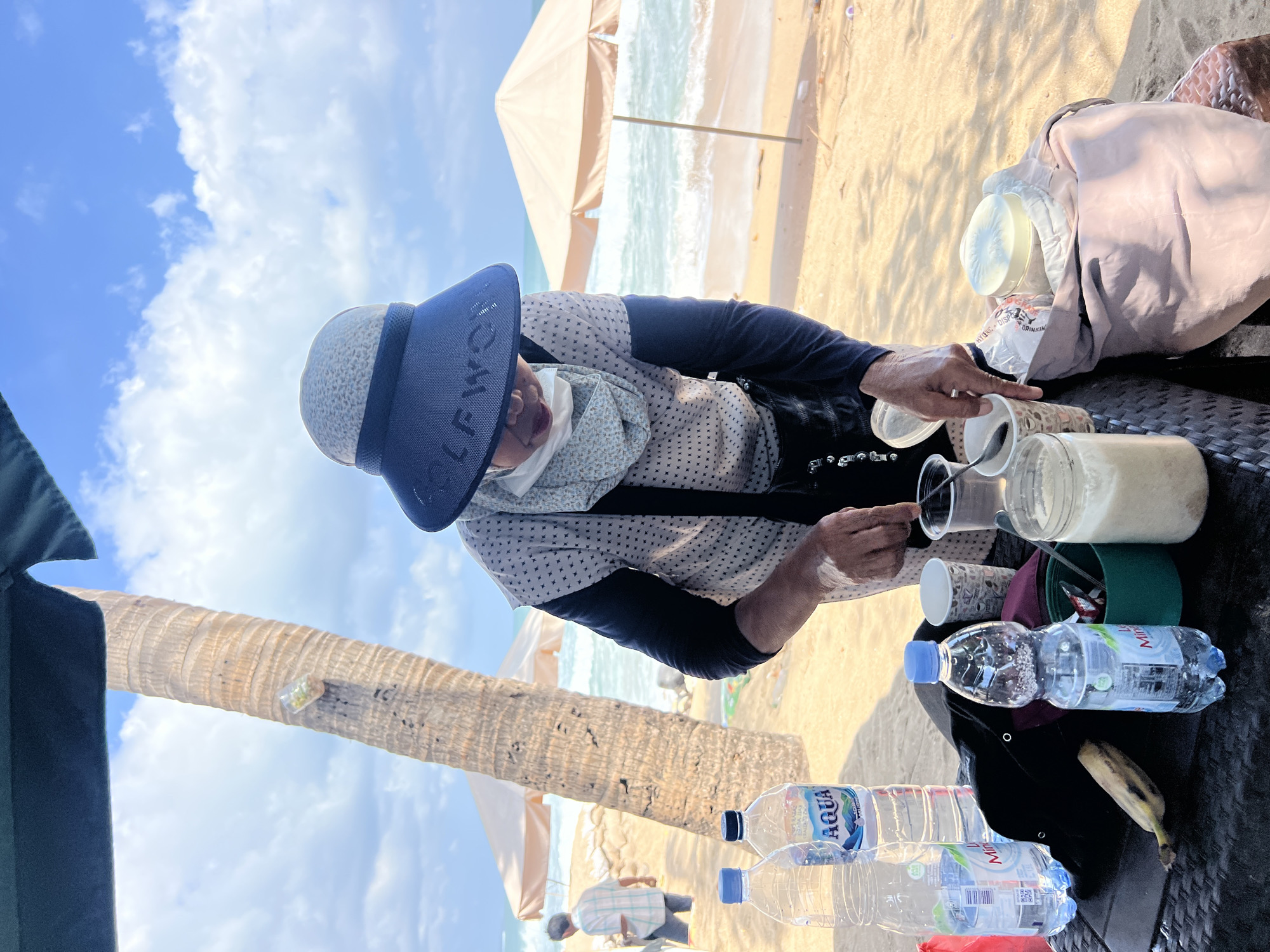 A person wearing a wide-brimmed hat and a face mask is standing on a beach in Kuta, Indonesia, preparing drinks. The person is in the process of scooping something from a cup with a spoon. In front of them is a table laden with various items, including several bottles of water, cups, a jar of white powder, and a banana. In the background, a palm tree stands tall, and the ocean waves crash on the sandy shore. Beach umbrellas are visible in the distance, suggesting a typical tourist spot. The sky is blue with scattered clouds, indicating a bright, sunny day. The overall scene depicts a person selling or preparing refreshments on a bustling beach.
