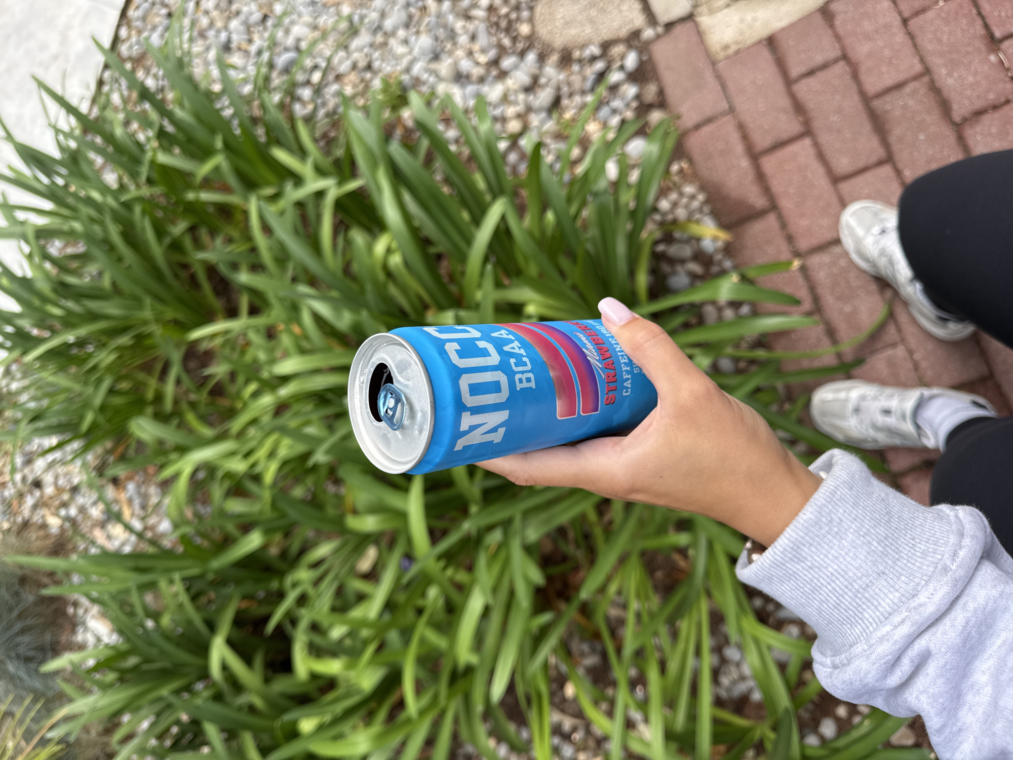 A woman holds an open blue can of NOCCO BCAA energy drink in her right hand. The setting appears to be outdoors near a garden with lush green foliage, adjacent to a brick pathway. The woman is wearing black leggings, white sneakers, and a light grey sweatshirt with the cuff visible. The location is not possible to determine to be the city of Tivat, Montenegro from this image alone.