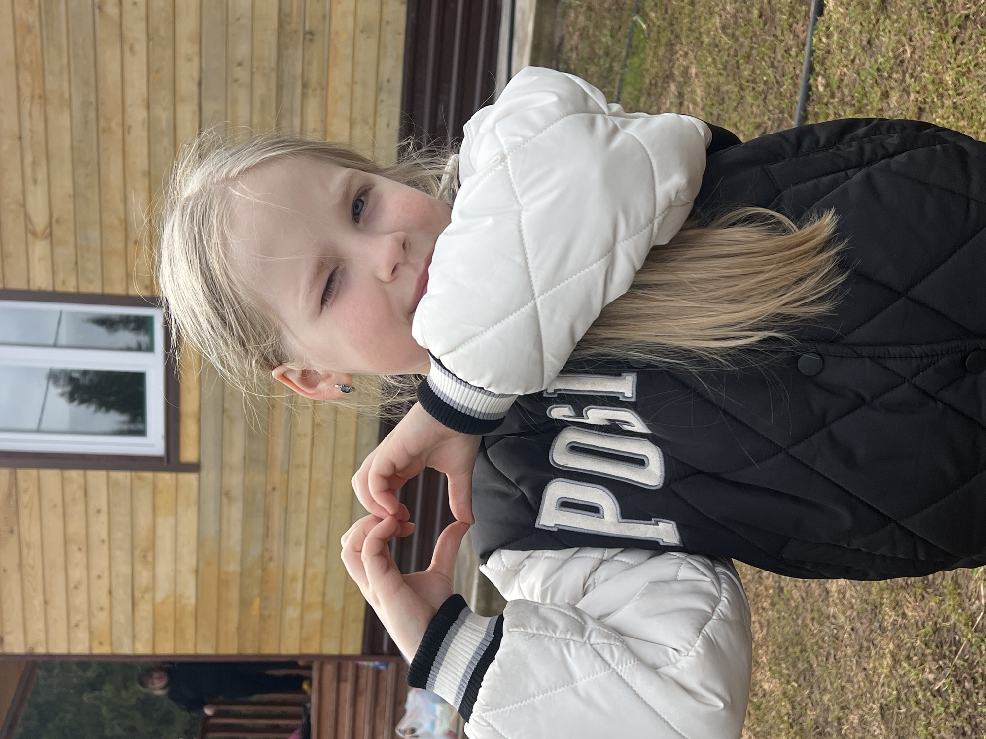 A young girl with blonde hair is playfully captured outdoors, rotated 90 degrees clockwise in the image. She is winking with her left eye and forming a heart shape with both hands, positioned in front of her chest. Her long, straight blonde hair cascades over her shoulders.
She is dressed in a quilted jacket, featuring a black body with white sleeves. The cuffs of her sleeves are ribbed with distinct black, white, and grey stripes. Partially visible text on the black section of her jacket appears to spell out "POST" or a similar word.
The background consists of a wooden building, likely a house or cabin, constructed with light-colored horizontal planks. A window with a white frame is visible on the building's side. The ground surrounding the structure appears to be sparse grass or dirt, suggesting a natural outdoor setting. The scene is illuminated by bright daylight, indicating good weather. The overall mood conveyed by the girl's expression and gesture is cheerful and loving. The location context is Bol'shakino, Russia.