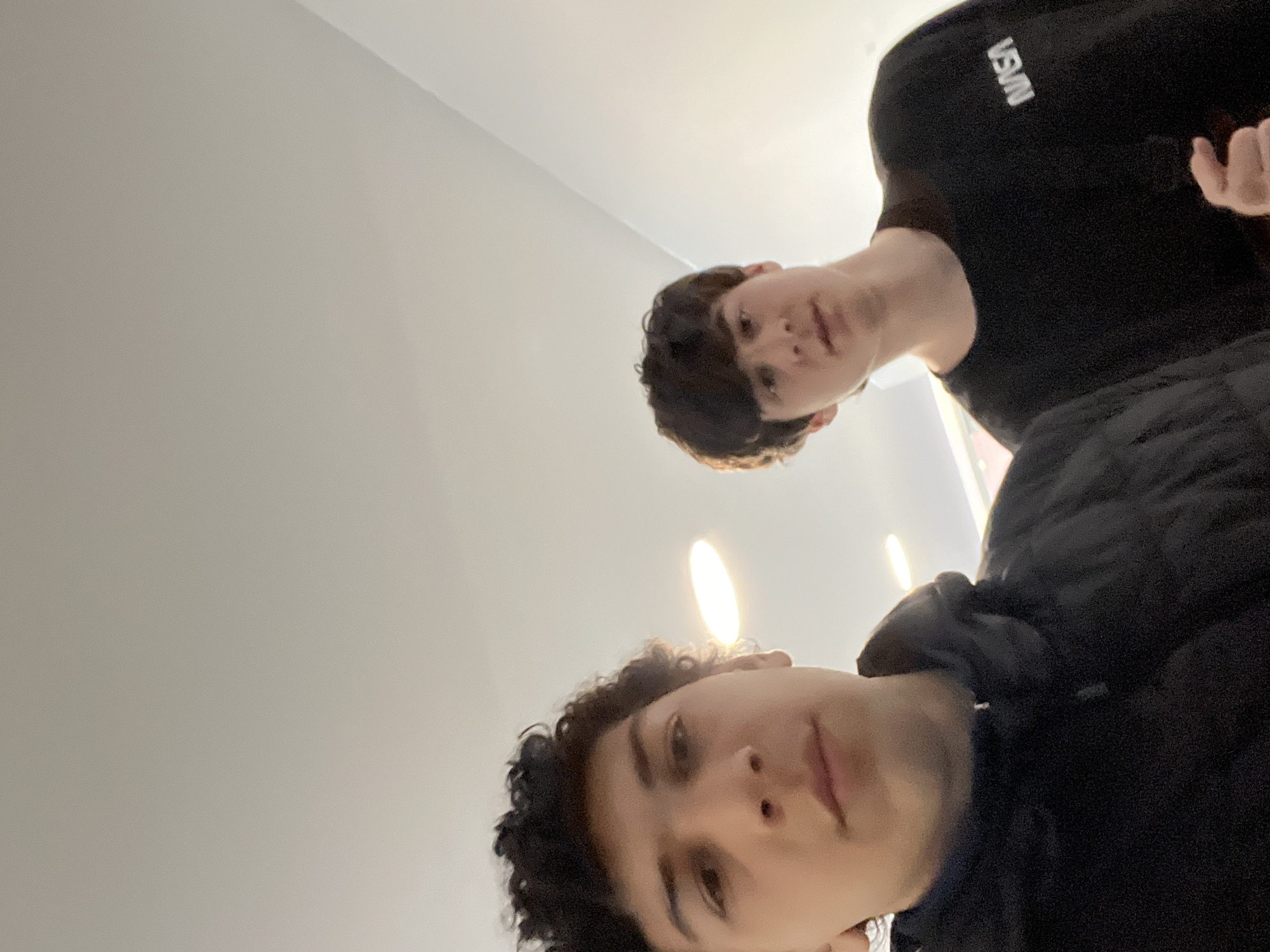 The image is a selfie taken from a low angle, showing two young men indoors. The man closer to the camera has curly dark hair and is wearing a black puffy jacket. The other man has straight dark hair and is wearing a black t-shirt with the word "NASA" in white lettering. The background is a plain white wall with ceiling lights. There is no information indicating the location is in Schiltigheim, France.
