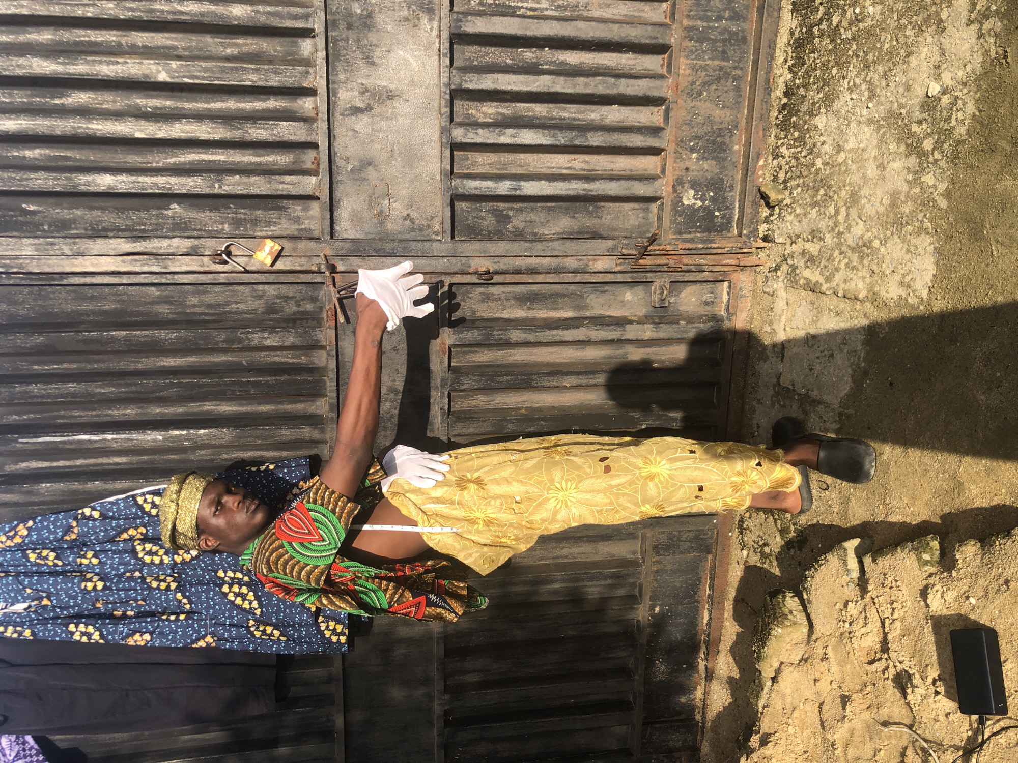 In Ado, Nigeria, a young Black male stands in front of a weathered, black corrugated metal gate. He is wearing a gold head wrap, a colorful patterned shirt with green, red, and yellow accents, and a flowing yellow skirt with intricate floral cutouts. White gloves are on both of his hands. In his right hand, he holds a white measuring tape, which extends down his torso. His left hand is slightly extended, also wearing a white glove. The ground in front of the gate is a mix of sand and concrete, with some scattered rocks and debris. To the left, a black electronic device with a wire is visible on the ground. Behind the man, fabric is draped, including a blue cloth with a yellow floral pattern and a black garment. The sunlight casts a shadow of the man and a nearby rock onto the ground. The gate has a rusty padlock securing it.