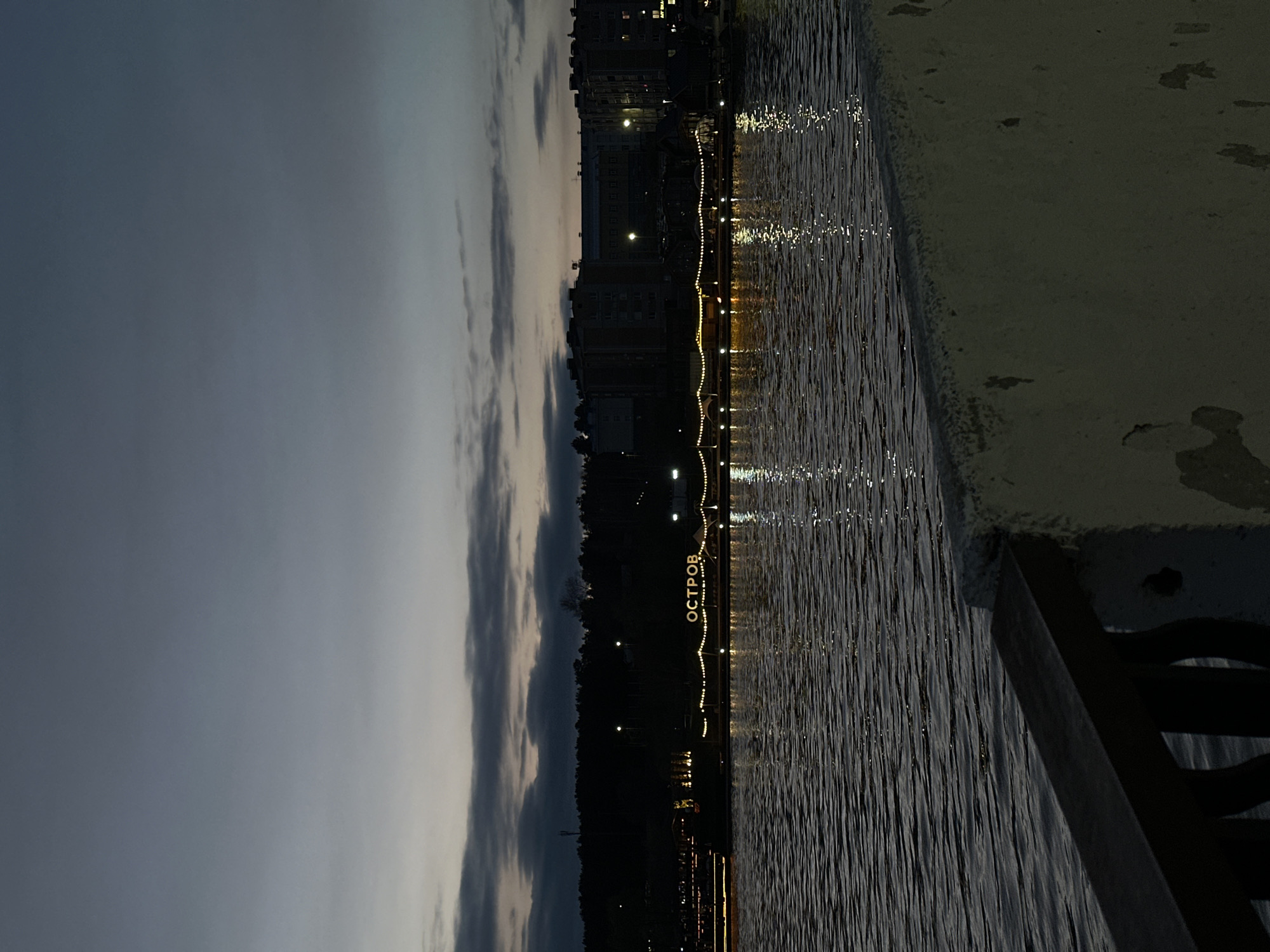 A nighttime, rotated, landscape-oriented photograph shows the embankment of Votkinsk, Russia, at night. The sky is a gradient of grays and blues with streaks of clouds. A body of water with reflected lights from the city dominates the center. Along the water’s edge is a series of illuminated buildings and trees. The word "ОСТРОВ" (OSTROV) is spelled out with lights along the shore. A concrete edge runs along the right side of the frame. A dark railing is visible in the lower right corner.