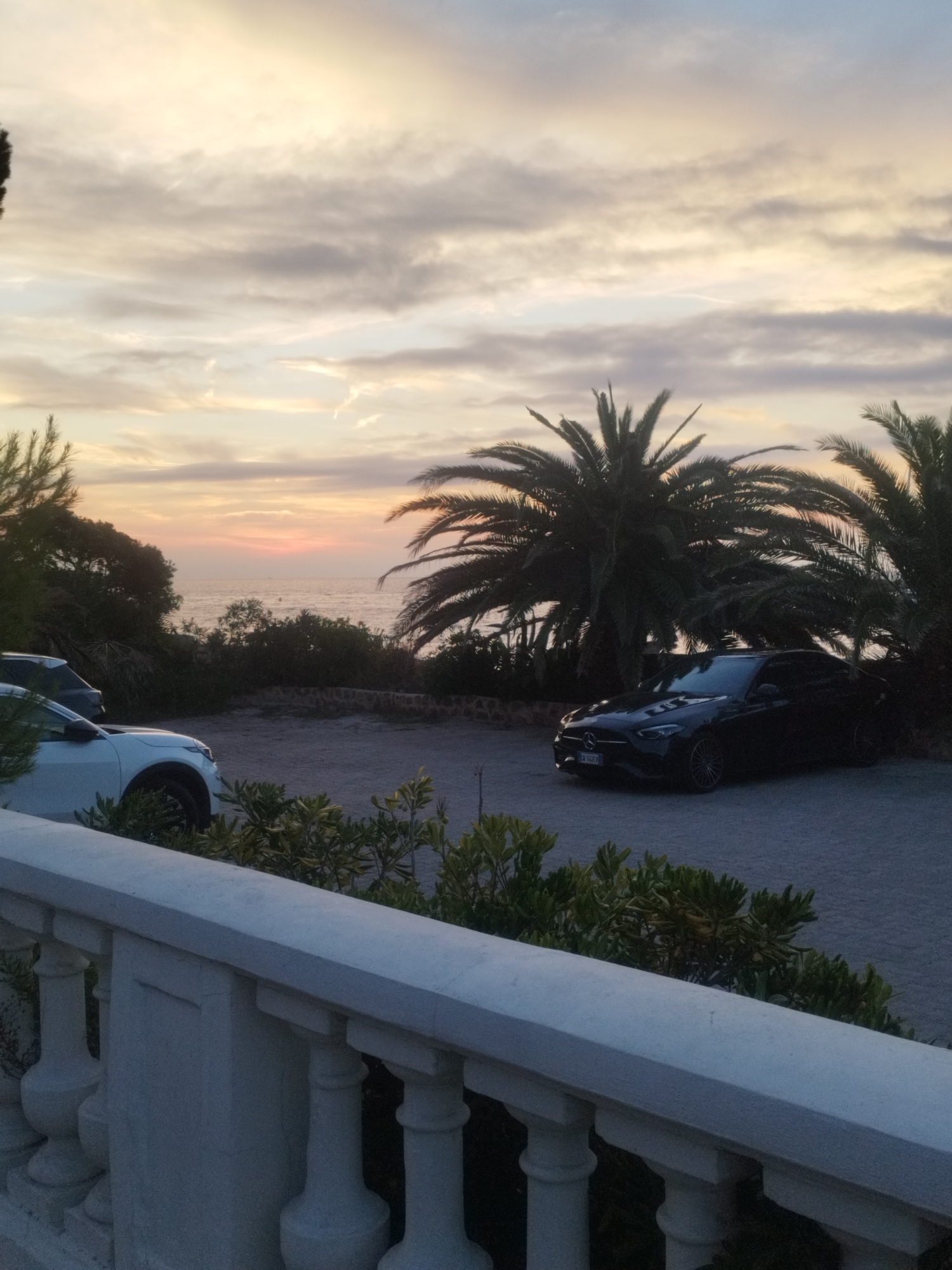 This is a slightly angled, medium shot of a coastal scene at dusk in Antibes, France, featuring two parked cars and lush vegetation. In the foreground, a white balustrade with decorative pillars lines the bottom of the frame. Beyond it, a white car is parked facing left, partially obscured by some greenery. To the right of the white car, a dark-colored sedan is parked facing right. The middle ground reveals a gravelly area with more foliage, including palm trees, and a low stone wall. The background showcases the calm sea stretching to the horizon, under a sky filled with soft, wispy clouds illuminated by the fading sunlight of dusk, which casts a warm, orange glow. The overall ambiance is peaceful and serene, suggestive of a residential or resort setting.