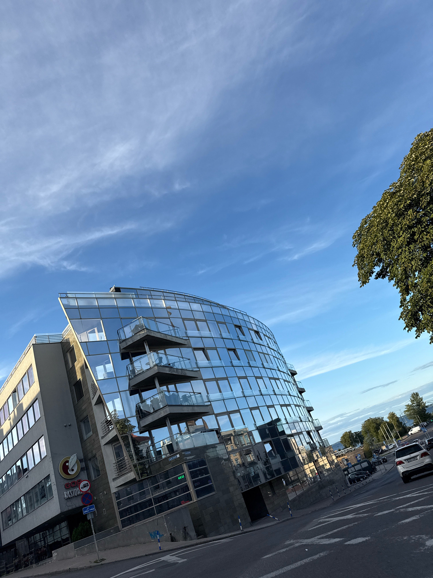 The image shows a street scene in Tallinn, Estonia, at an oblique angle, with a modern, curved glass building dominating the left side. The building has multiple floors with balconies and is characterized by its reflective glass facade. To the left of the curved building, a more traditional, rectangular building with windows is visible. A sign on the rectangular building reads "Gop Restora". Below the buildings, a street with white markings and a sidewalk are present. Several cars are visible on the street, including a white SUV in the foreground on the right. A large tree with green foliage is positioned in the upper right corner of the frame, partially obscuring the sky. The sky is blue with thin, wispy clouds. The overall impression is that of an urban environment during daylight hours.
