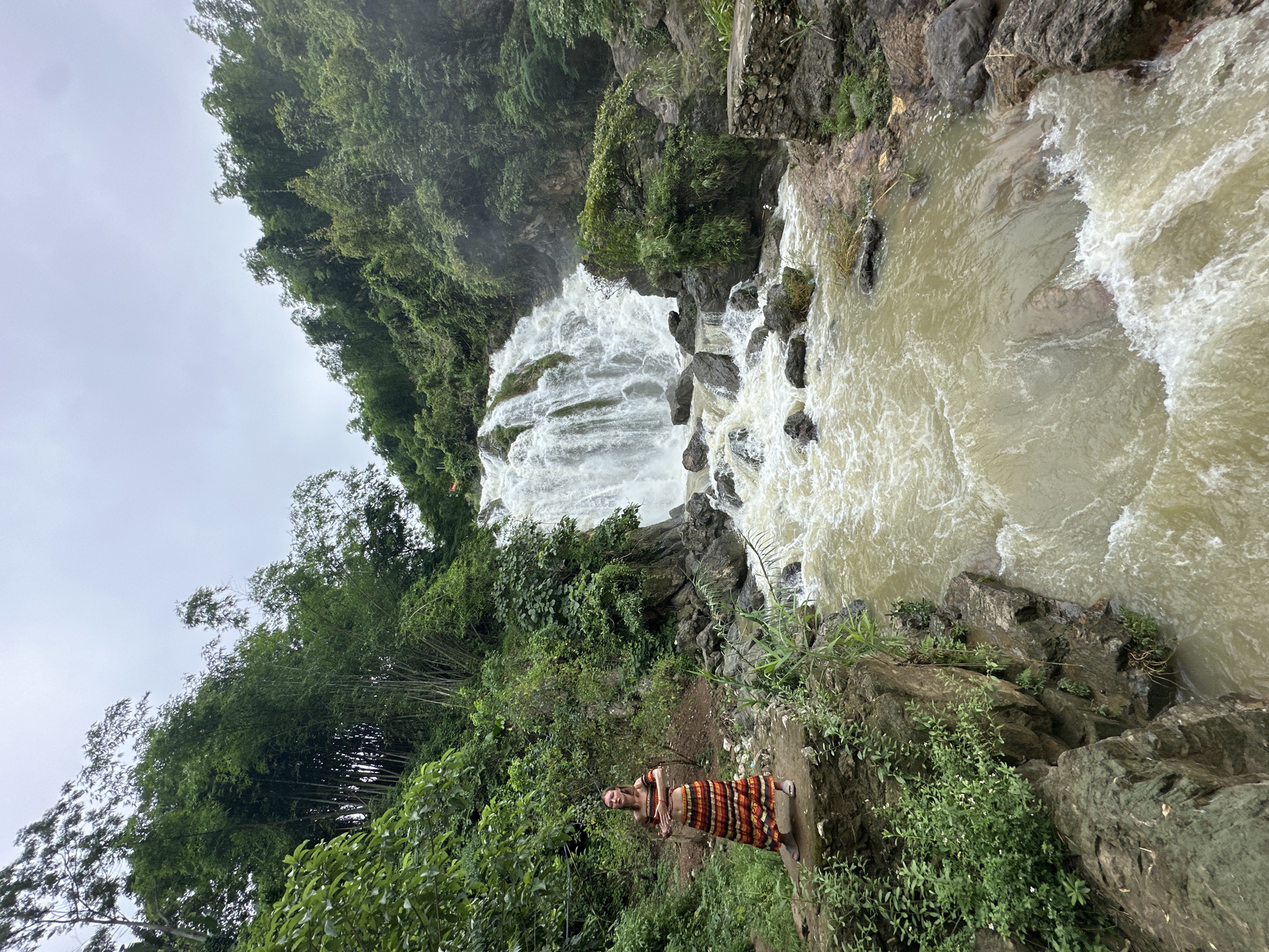 A woman stands on a rocky outcrop to the left of a powerful waterfall in Bản Phúc, Vietnam. The waterfall cascades down a steep, rocky incline, with a frothy, brown river rushing below. The woman, wearing a colorful striped skirt and a matching off-the-shoulder top, has her arms outstretched as if to embrace the natural scene. Lush green vegetation, including bamboo trees, surrounds the waterfall, creating a dense, tropical atmosphere. The sky is overcast, suggesting a humid, possibly rainy day. The overall impression is one of a serene and majestic natural landscape.