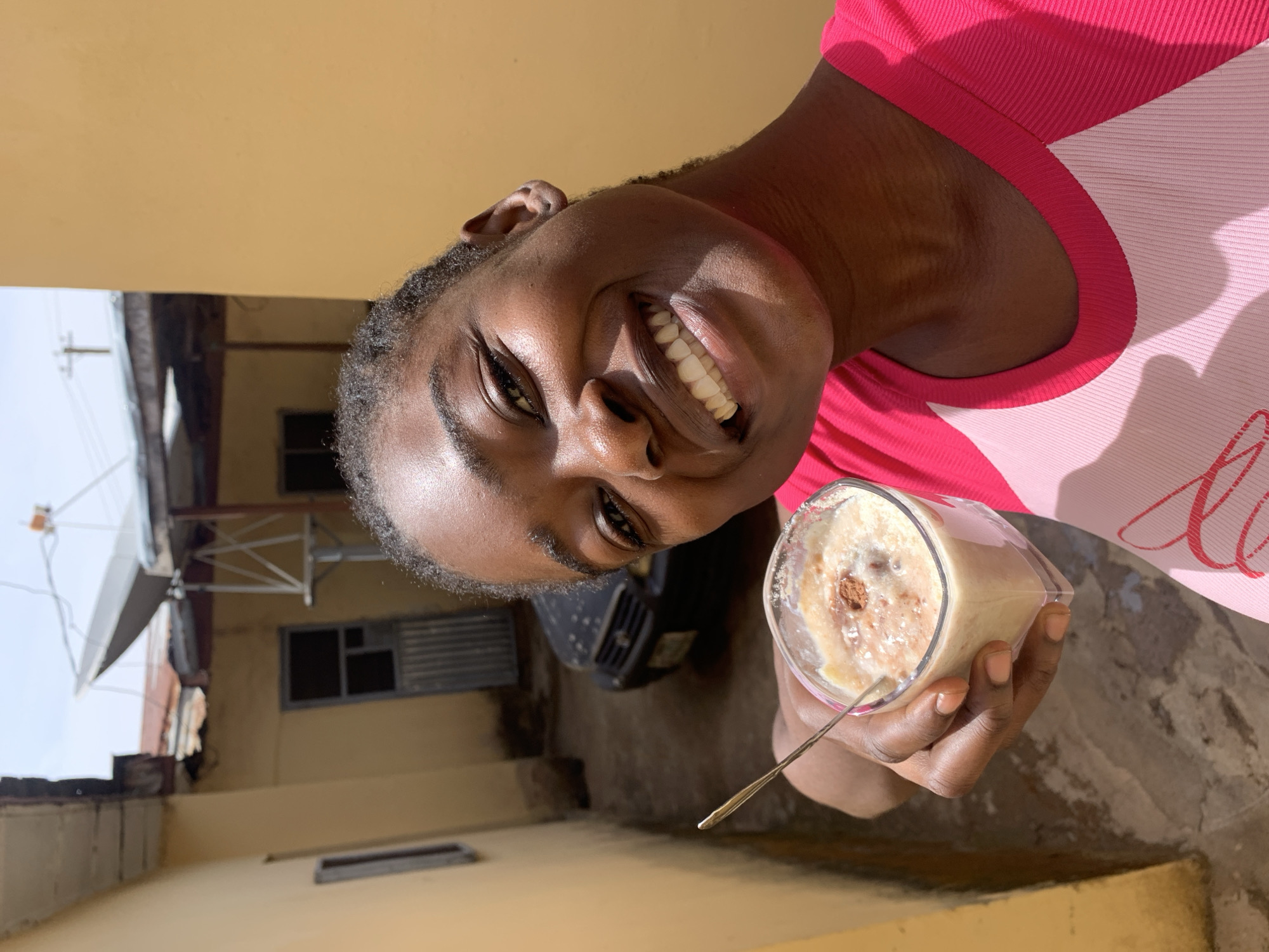 A medium shot of a Black woman holding a glass of a creamy, brown-topped beverage in a courtyard in Rafin Par, Nigeria. She is smiling broadly, wearing a pink shirt. The background shows a courtyard with cement flooring and tan walls. A metal satellite dish is visible on a nearby building. A black vehicle is partially visible behind her.