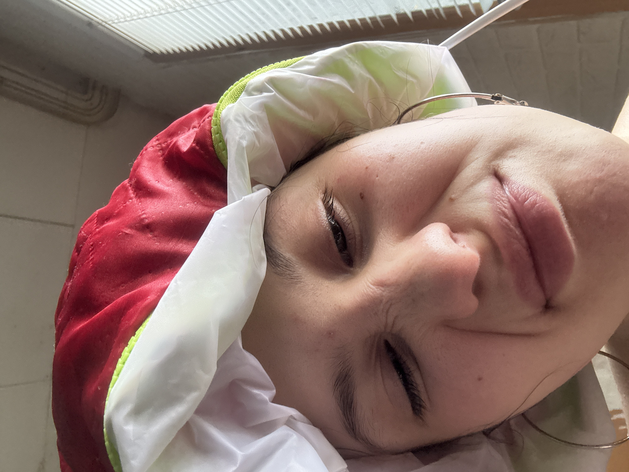 A close-up, angled shot features a person wearing a red and white shower cap. The person's face is angled toward the camera, with eyes mostly closed. The background consists of a textured ceiling. The location is likely not in Фоки, Russia as the image does not provide any relevant details.