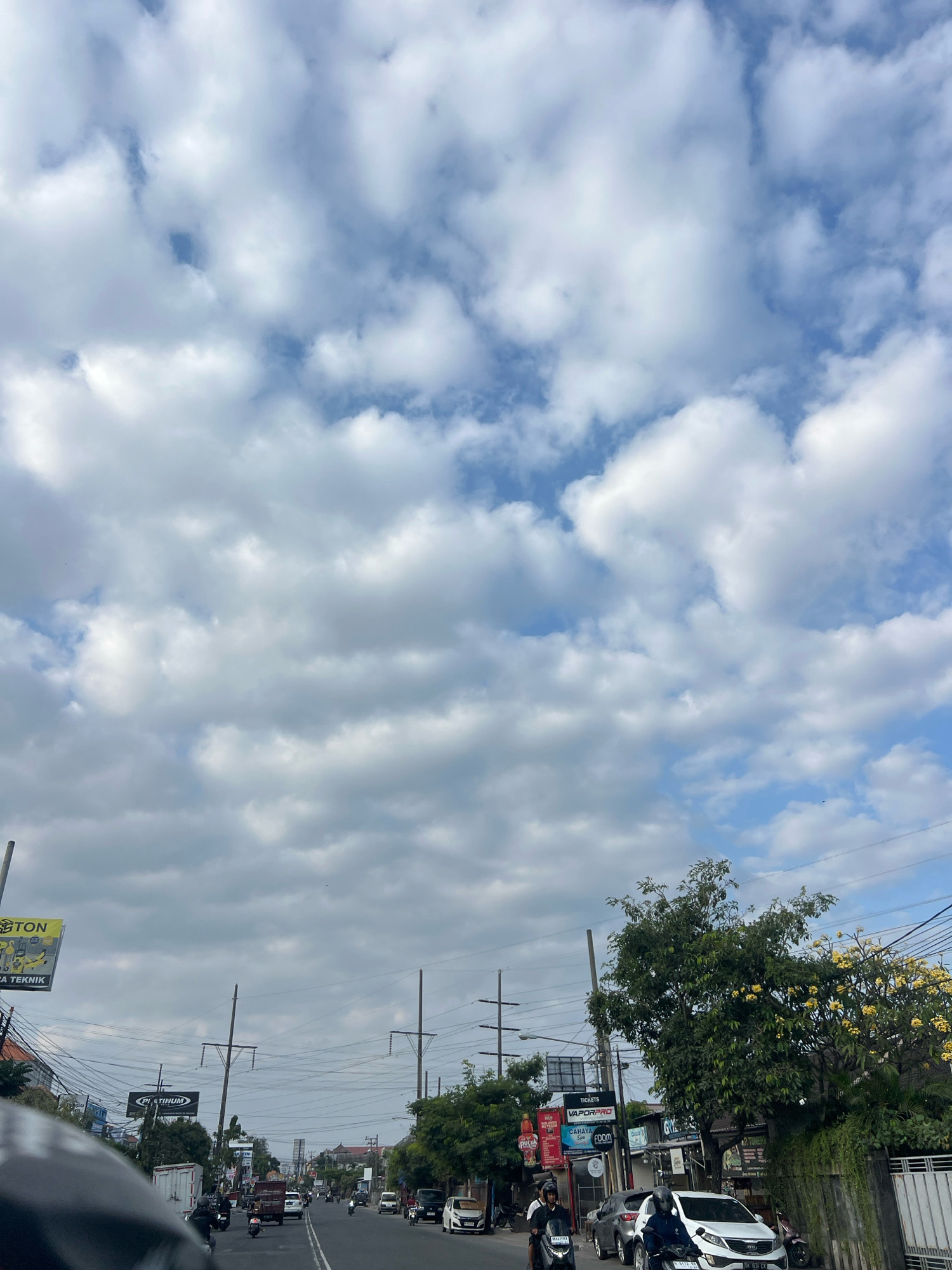 This is a street-level, vertical, medium-shot image of a busy road in Denpasar, Indonesia, under a partly cloudy sky. The road is asphalt with visible lane markings and is lined with businesses, trees, and utility poles. Traffic includes several motorcycles, cars, and a truck. Some of the visible businesses have signage, including "TON," "PLATINUM," and "TICKETS VAPORPRO." A large tree with yellow flowers is prominent on the right side of the image. The overall atmosphere is that of an active urban environment.