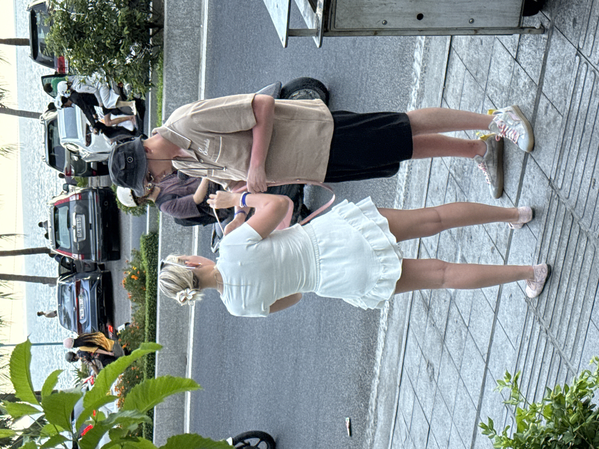 In Nha Trang Ward, Vietnam, a street scene unfolds with two individuals, a woman and a man, interacting near a sidewalk. The woman, viewed from behind, has blonde hair styled in a bun with a floral accessory and wears a white short-sleeved top and a ruffled white skirt. She appears to be holding something out towards the man. The man, facing the woman, is wearing a light brown short-sleeved collared shirt with "Sandals" embroidered on it, black shorts, a black bucket hat, and sunglasses. His arms are visible, with redness suggesting sunburn. He is wearing light-colored sneakers with pink laces. Behind them, a street is visible with parked cars, including a dark SUV and a lighter sedan, and several people on motorbikes. In the background, palm trees and the ocean are present. The foreground shows a tiled sidewalk and some greenery.
