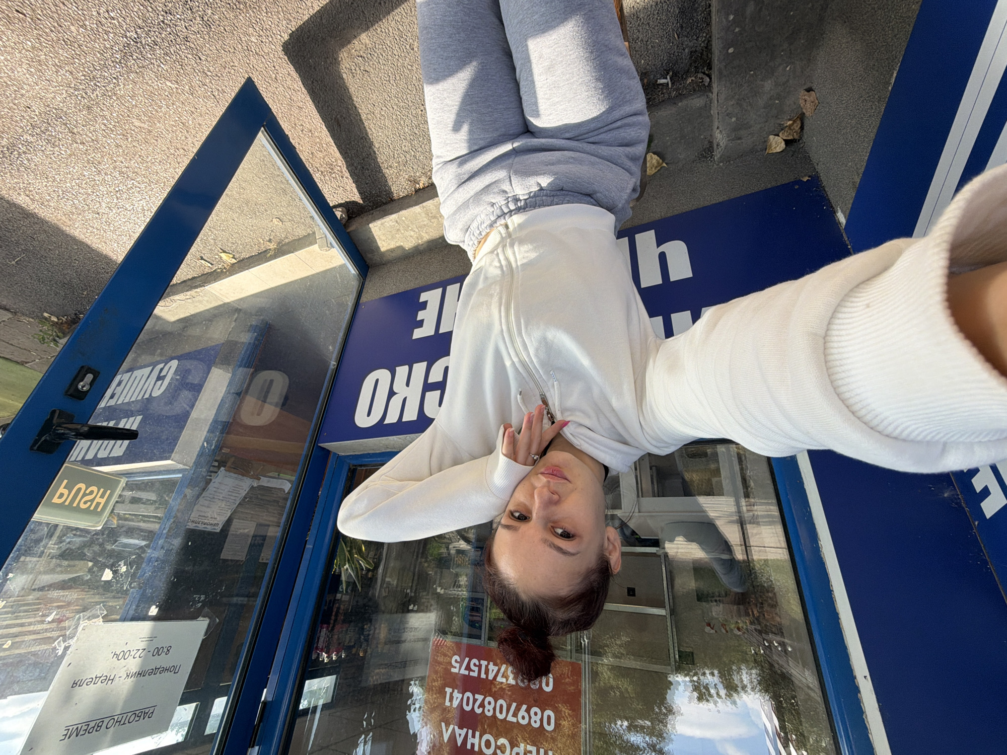 A young woman with her hair pulled up in a bun takes a selfie in front of a glass-enclosed structure. She is wearing a white zip-up hoodie and gray sweatpants. The glass displays various signs, some in Cyrillic script. One sign reads "РАБОТНО ВРЕМЕ" (Working Hours) and lists hours from 8:00 to 22:00 daily. Another sign has a phone number, "0897082041". The structure appears to be a kiosk or small shop. The background shows trees and a cloudy sky, suggesting an outdoor urban environment. The kiosk's blue frame and the gray concrete steps indicate it is situated in Sofia, Bulgaria.