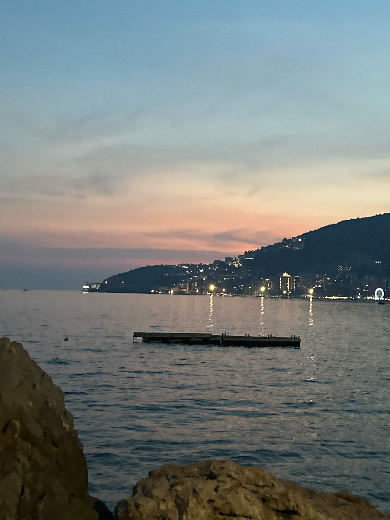 The image shows a twilight view of the Budva Riviera coastline in Montenegro. The sky exhibits a gradient of pale blue at the top transitioning to soft pink and orange hues near the horizon, characteristic of a sunset or sunrise. The calm sea occupies the majority of the lower half of the image, with gentle ripples reflecting the dim light and scattered city lights. In the foreground, on the left and bottom right, are large, rough rocks, suggesting a rocky shoreline.
Further out in the water, a dark, rectangular platform or pier with small vertical posts is visible. In the mid-ground and background, the coastline is lined with numerous lights from buildings that ascend a dark, mountainous hillside. The lights create a sparkling effect against the darkened landscape, indicating a populated area. A prominent Ferris wheel, lit with white lights, can be seen on the right side of the image, towards the top of the illuminated hillside. In the far left distance, a small boat is faintly visible on the water. The overall atmosphere is serene and peaceful, capturing the transition from day to night along the Adriatic coast.