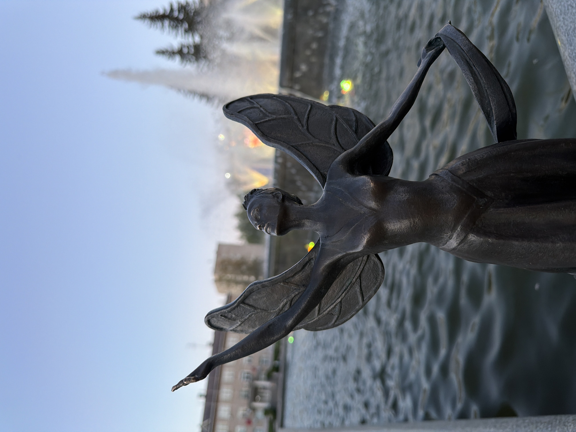 The image shows a bronze statue of a winged woman, possibly an angel or fairy, with one arm outstretched and the other holding a ribbon or cloth above her head. The statue is positioned in front of a fountain with multiple jets of water shooting upwards. Buildings are visible in the background, suggesting an urban setting. The sky is a pale blue, indicating either early morning or late evening. The statue is the focal point, with the fountain and buildings serving as background elements. The photo was taken in the city of Chaikovsky, Russia.