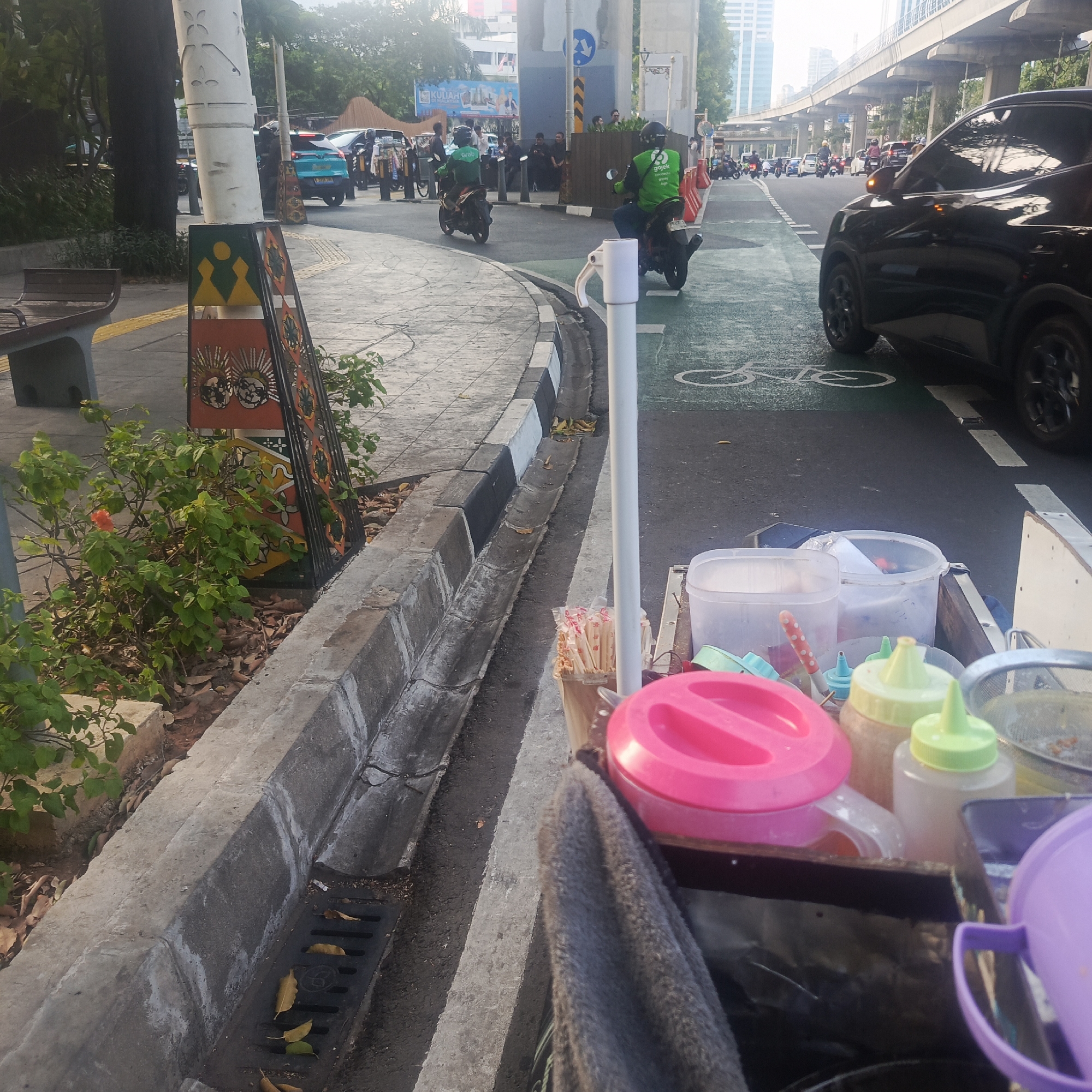 The image depicts a street scene in South Jakarta, Indonesia, likely during daylight hours. In the foreground, a close-up view shows a food cart laden with various containers and utensils, including a large pink pitcher, several bottles of condiments with green and yellow caps, a bundle of chopsticks, and a metal sieve. A gray cloth is draped over part of the cart. The cart is positioned next to a concrete drain and a sidewalk. Beyond the cart, a road is visible with a green-painted bike lane marked with a white bicycle symbol. Several vehicles, including cars and motorcycles, are present on the road, with some drivers wearing green vests, suggesting they may be delivery personnel. A black car is prominently in the right side of the frame. Further in the background, elevated highway structures and buildings are visible, characteristic of an urban environment. Lush green foliage and trees line the sidewalk and roadside, with one small orange flower visible on a bush. The scene appears to be a busy street with ongoing traffic and pedestrian activity, suggesting a commercial or residential area within South Jakarta.