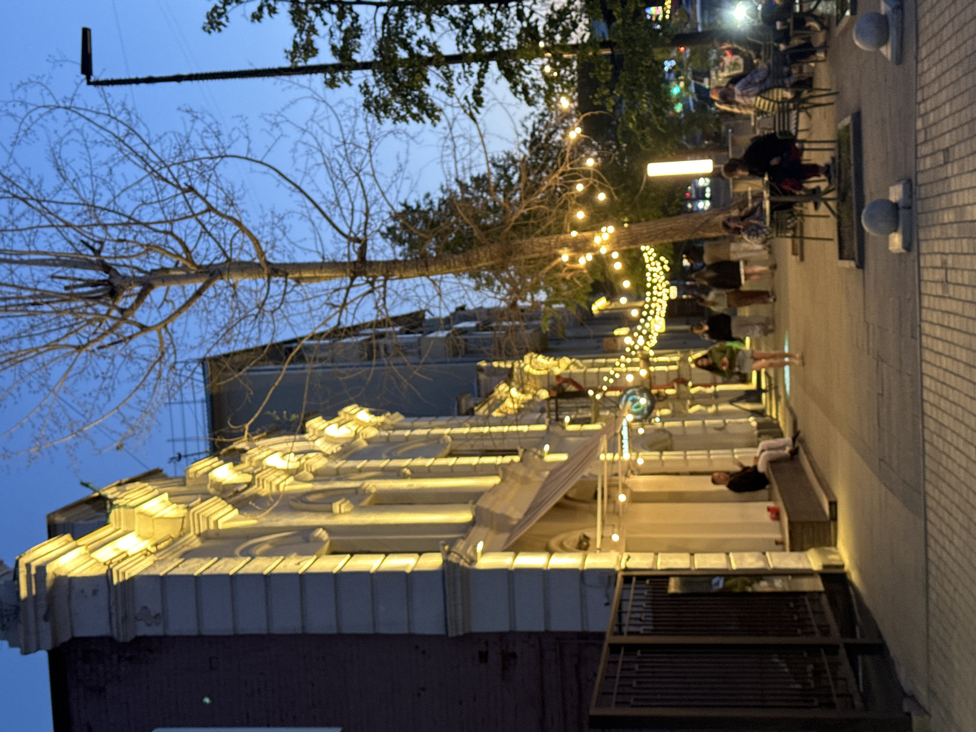 The image depicts a street scene at dusk in Krasnoyarsk, Russia. On the left, a building with ornate yellow lighting and classical architectural features is visible. A dark metal fence stands in the foreground, to the left of a sidewalk. A person is seated on a bench along the building's facade.
The street itself is paved with brick-like patterns. Further down the sidewalk, several people are walking, and groups are seated at tables outside what appears to be a cafe, illuminated by strings of warm fairy lights. Trees, one bare and branchy, and others with foliage, are present, with a tall street lamp to the right. The sky is a deep blue, indicating the transition from day to night. The overall atmosphere is urban and social, with an emphasis on outdoor dining and evening ambiance.