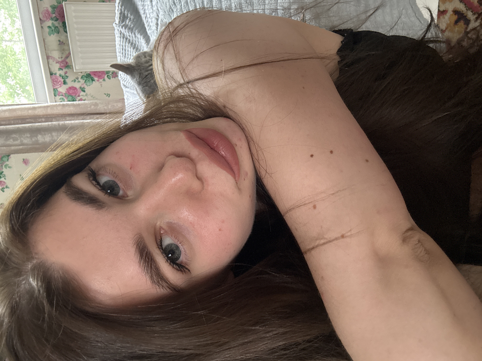A young woman is seen close-up in an indoor setting. She has fair skin, dark eyebrows, and light-colored eyes. Her long, brown hair is disheveled around her face and arm. She is wearing a dark-colored top. The background includes a window with floral wallpaper, a radiator, a partially visible gray cat, and a bed with gray bedding. A rug with a geometric pattern is also visible. Location cannot be determined without further information.