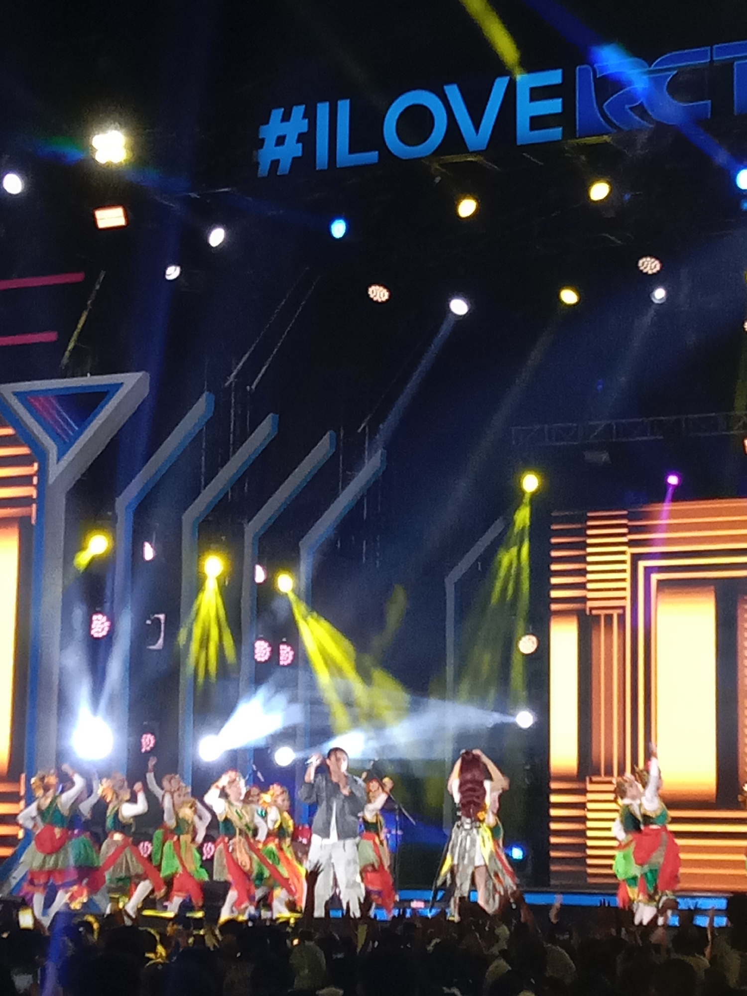 An outdoor stage performance is underway, featuring a group of female dancers in colorful traditional Indonesian attire and a male singer. The stage is illuminated by bright spotlights and a large LED screen displaying the hashtag "#ILOVERCTI". The dancers are in motion, performing choreographed routines, while the singer stands at the microphone, engaging with the audience. The scene suggests a vibrant cultural event taking place in Kendal, Indonesia, potentially a festival or concert. The audience is visible in the foreground, their hands raised in appreciation. The overall atmosphere appears energetic and festive.
