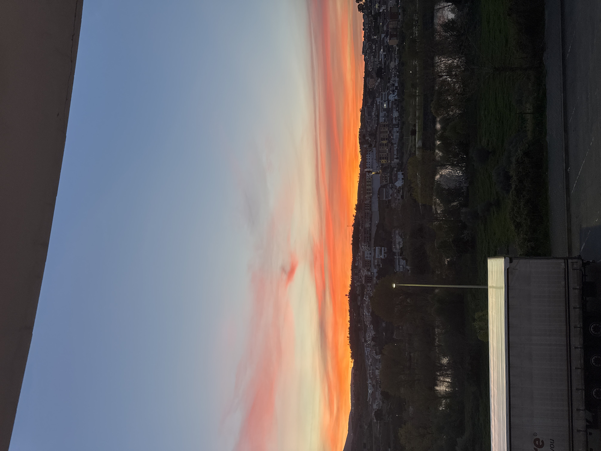 The image shows a sunset view in Mirandela, Portugal. The sky transitions from blue to orange and red hues, with scattered clouds. Below the skyline, a dense residential area with houses and buildings is visible, blending into the landscape. A long, vertical lamp post is present, casting light in its immediate vicinity. To the right, there's a grassy area and a paved surface, with part of a white truck parked nearby. The left side of the frame shows a fragment of a building's corner.