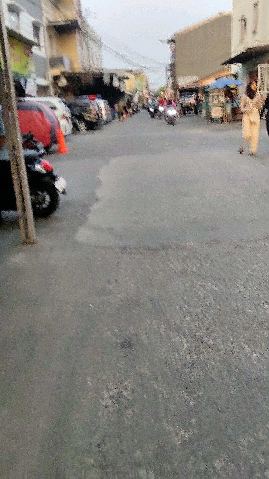 The image depicts a street scene in Mekarmukti, Indonesia, likely during daylight hours. In the foreground, the asphalt road is visible with some areas of dampness or discoloration. To the left, a portion of a black motorcycle is parked next to a vertical metal pole. Further down the street, several vehicles are parked along the roadside, including what appears to be a black SUV and white cars. A red object, possibly a tarp or cover, is draped over a vehicle or object. Orange traffic cones are also present.
The middle ground shows a street with several motorcycles and people. Some motorcycles appear to be in motion, with their headlights illuminated. Pedestrians are walking along the street. On the right side of the street, there are shops or stalls, with awnings and umbrellas providing shade. One stall has what looks like a food cart. A woman wearing a headscarf and light-colored clothing is walking on the right side of the street.
In the background, buildings line both sides of the street. The buildings exhibit a range of architectural styles and colors, with some appearing older and others more modern. Electrical wires are strung overhead. The sky is overcast or hazy. The overall impression is of a busy urban street environment.