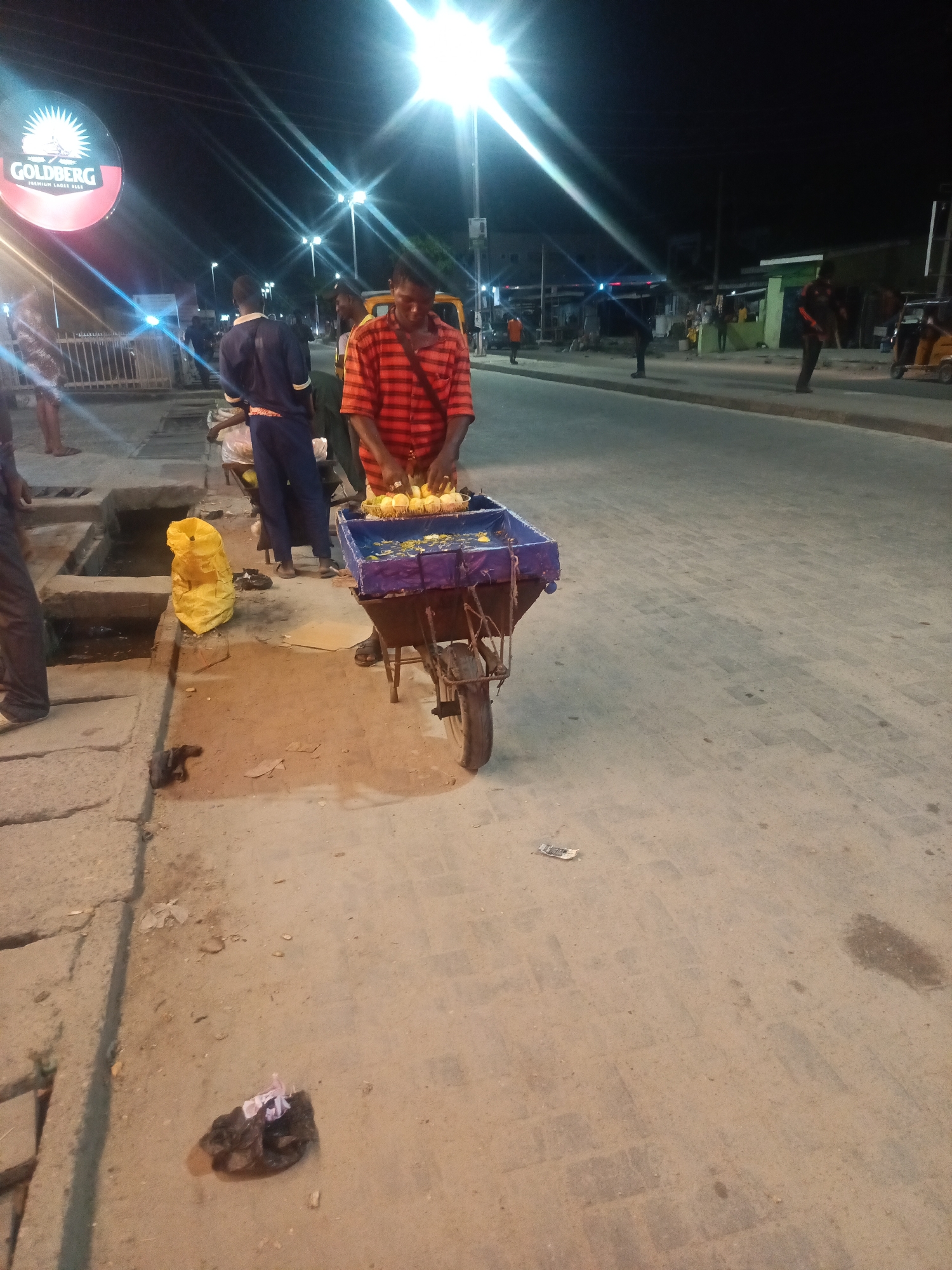 At night in Ado, Nigeria, a man in a red and orange striped shirt stands behind a wheelbarrow filled with corn. The wheelbarrow's tray is purple and decorated with yellow and blue flowers. He is using his hands to arrange the corn. To his left, a bright yellow plastic bag rests on the ground near a concrete gutter. Behind the man, other people are visible, including one in a blue uniform and another wearing an orange shirt. In the background, streetlights illuminate the paved road and sidewalks, casting long shadows. A large circular sign with the word "GOLDBERG" and an image of a sunburst is prominently displayed on the left side of the scene, indicating a commercial area.