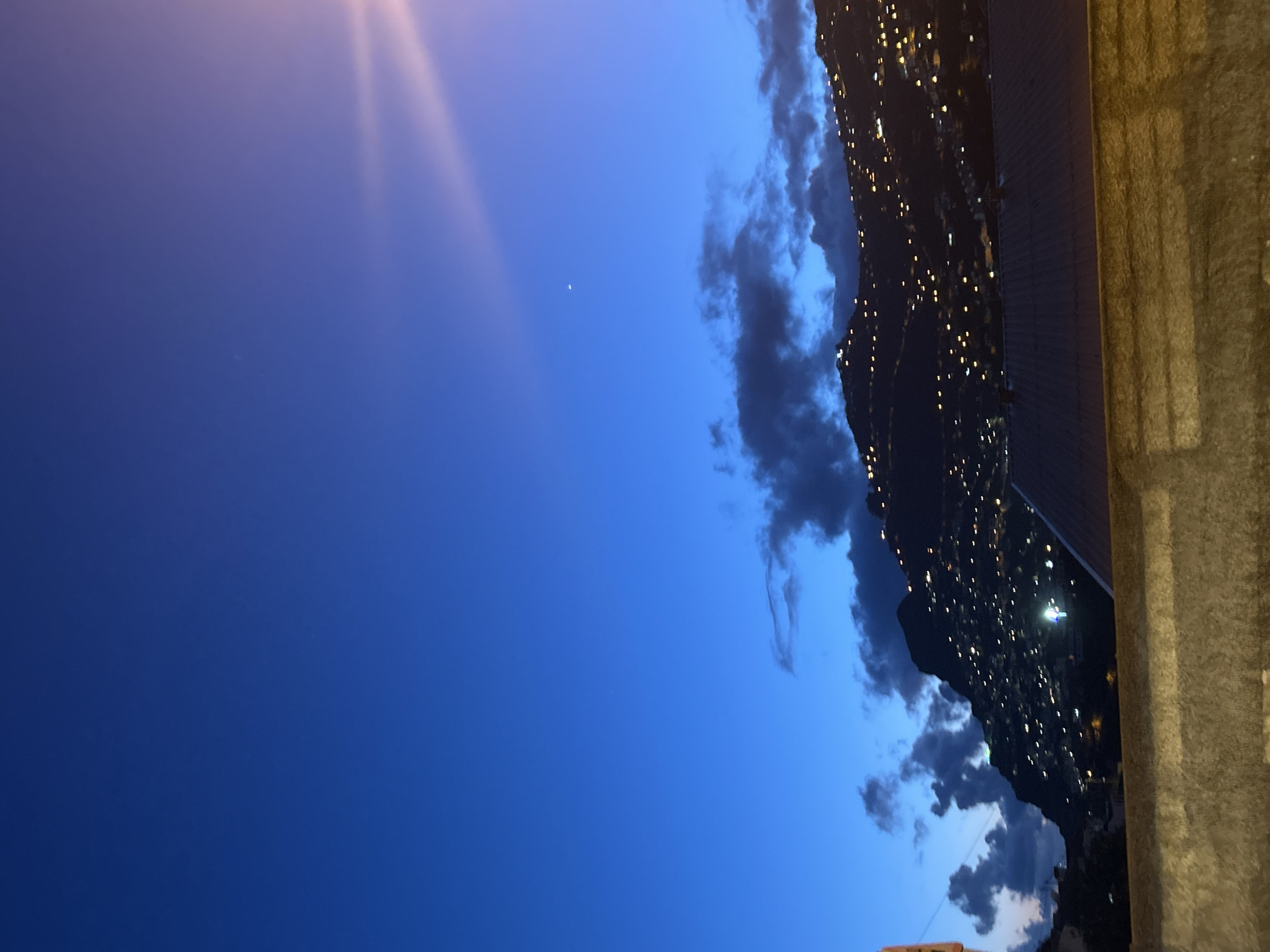 The image shows an evening view of Câmara de Lobos, Portugal, from an elevated perspective. A predominantly deep blue sky fills the majority of the frame, with a visible light flare in the upper left corner and a faint crescent moon. Along the right side, a dark landmass is lined with numerous small, bright lights, suggesting a densely populated area or city illuminated at night. The edge of a concrete structure or wall is visible on the far right, providing the viewpoint. The landmass is partially obscured by dark clouds in the middle.