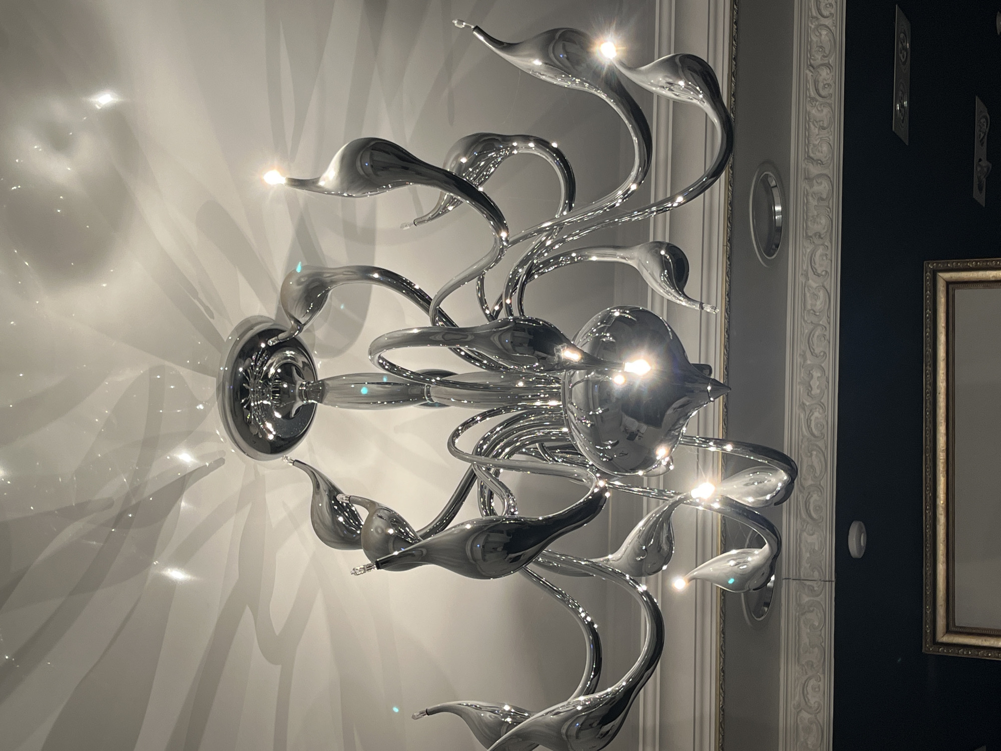 The scene depicts an interior space in Neftekamsk, Russia. A large, multi-armed chandelier, constructed from highly polished metallic elements with a curvilinear, organic design, is suspended centrally from a white ceiling. Numerous sinuous arms extend from a central base, each terminating in a reflective, teardrop-shaped component containing an exposed incandescent light bulb. Several bulbs are illuminated, casting bright points of light and producing distinct wavy shadows and glittering reflections across the white ceiling. Below the chandelier, a decorative white ornate cornice separates a light-colored upper wall section from a darker, possibly black or dark blue, lower wall. A partially visible, gold-framed object is present in the lower center of the frame. Circular recessed light fixtures are embedded in the upper wall, and a small, white, circular device, consistent with a smoke detector or sensor, is mounted on the darker wall. No individuals or actions are depicted.