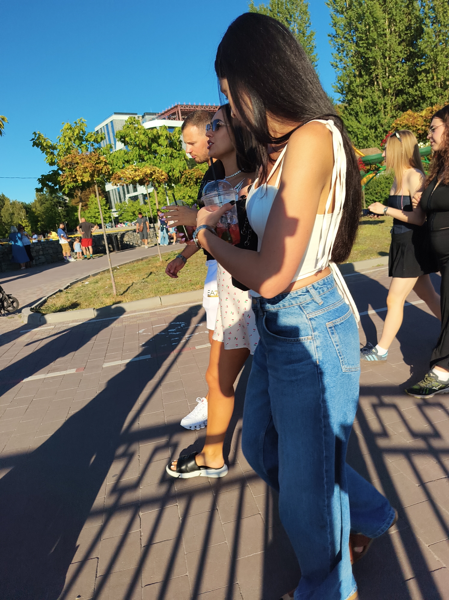 The image shows a group of people walking on a paved pathway on a sunny day. In the foreground, a woman with long dark hair is wearing a white top and blue jeans. To her left, a man with short brown hair and sunglasses is walking. Behind them, other individuals are visible walking and standing. The background features trees, buildings, and a clear blue sky. The scene appears to be a public area, possibly a park or a promenade. Given the context of Ternopil, Ukraine, this location could be the Ternopil Lake embankment or a similar recreational area.