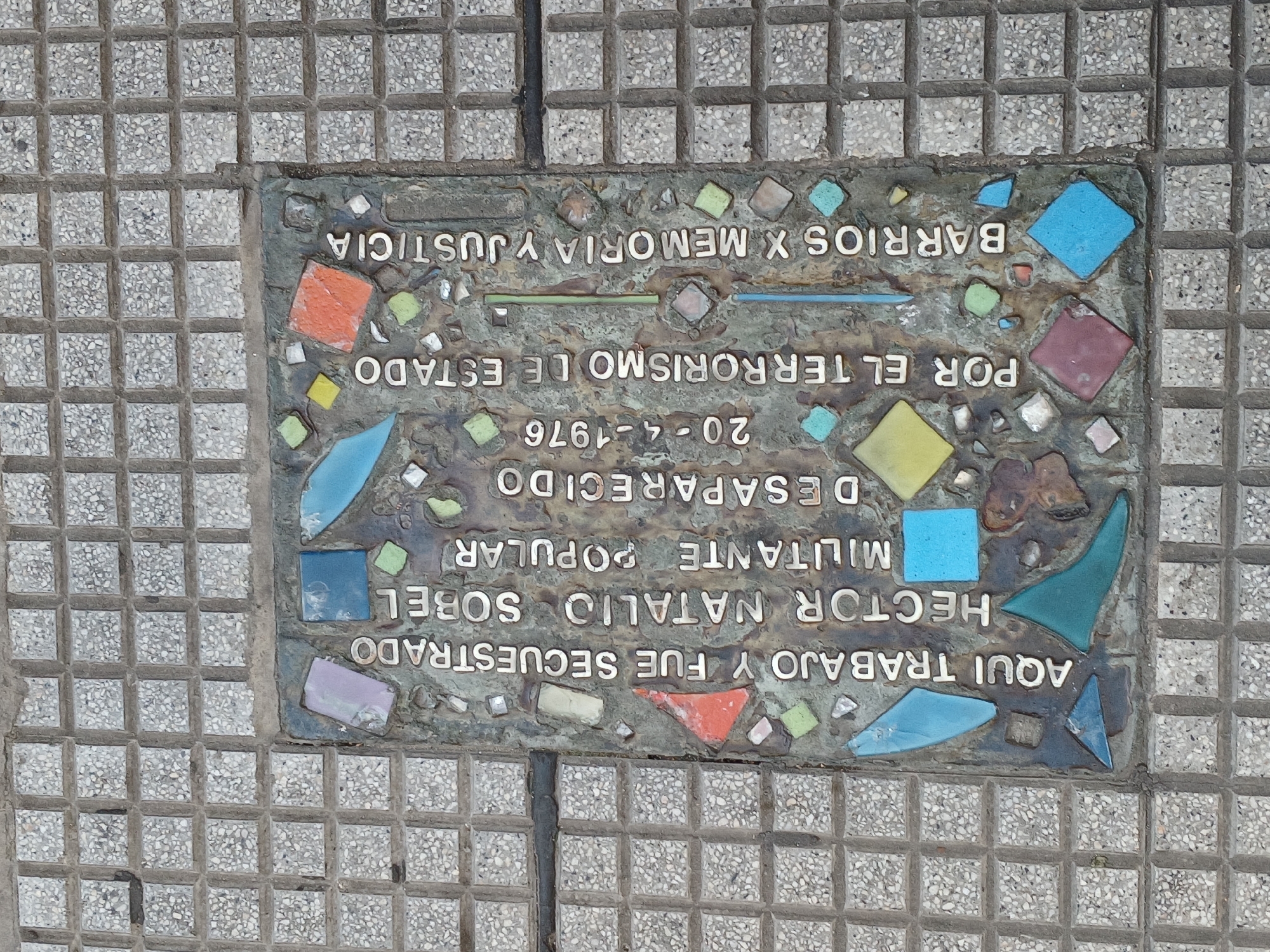 The image shows a commemorative plaque embedded in the sidewalk, likely in Buenos Aires, Argentina. The plaque is bronze and decorated with colorful tile fragments. Inscribed on it are the words: "AQUI TRABAJO Y FUE SECUESTRADO HECTOR NATALIO SOBEL, MILITANTE POPULAR DESAPARECIDO 20-4-1976 POR EL TERRORISMO DE ESTADO BARRIOS X MEMORIA Y JUSTICIA", meaning "Here worked and was kidnapped Hector Natalio Sobel, Popular Militant, Disappeared, 20-4-1976, By State Terrorism, Neighborhoods for Memory and Justice". The plaque is surrounded by interlocking square concrete pavers.