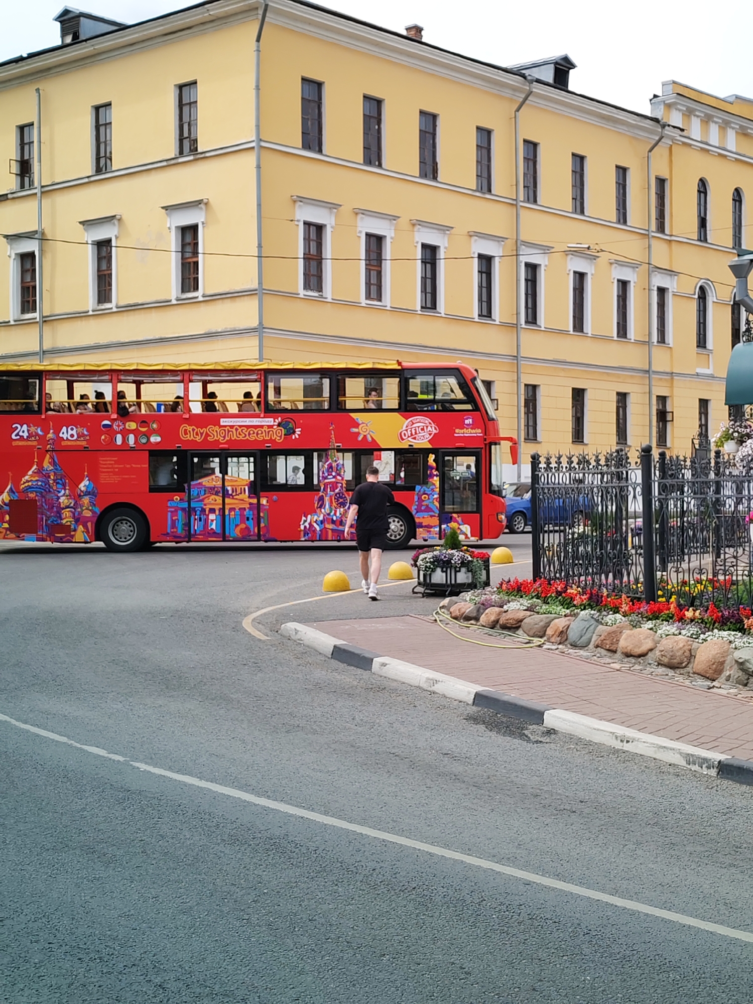 An eye-level, medium shot captures a bright yellow, multi-story building in the background. The building has numerous windows and ornate white trim around some of them. In the foreground, a red double-decker tour bus is parked on a paved road. The bus is adorned with colorful illustrations of what appear to be historical Russian landmarks. On the upper deck of the bus, passengers are visible. A lone man wearing a black t-shirt and shorts walks away from the bus towards the right of the frame. He is on a paved sidewalk that is bordered by a decorative garden with colorful flowers and large rocks. To the right of the garden, there is a black wrought-iron fence. The sky is overcast and a blue car is parked behind the tour bus, further down the road.