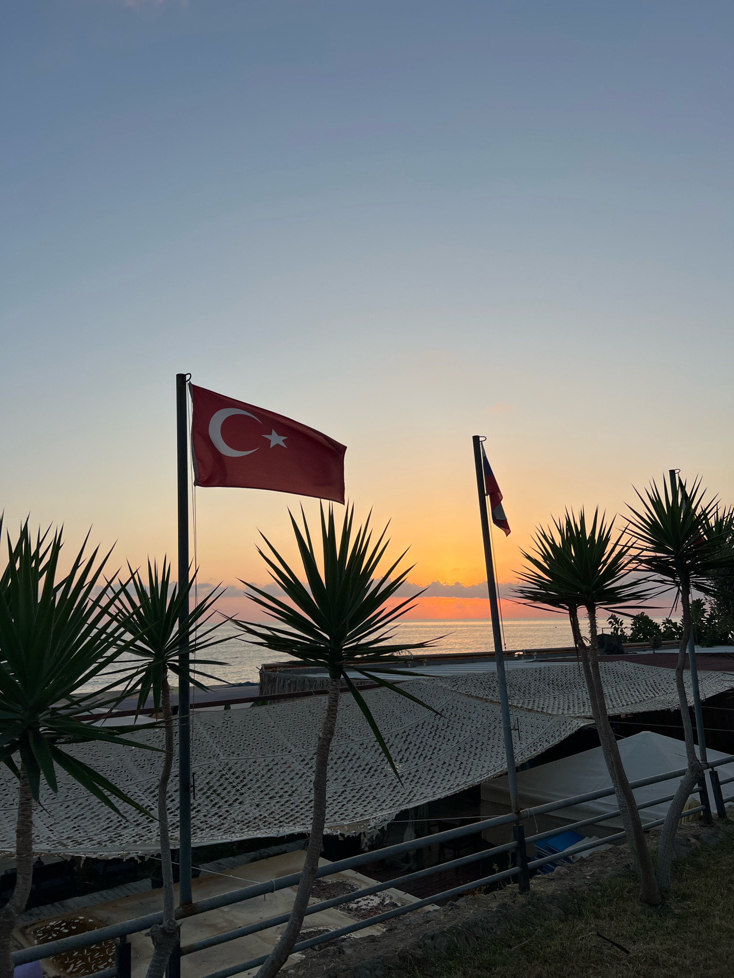 The image captures a serene sunset scene in Alanya, Turkey, likely from an elevated vantage point overlooking the Mediterranean Sea. In the foreground, several yucca plants with sharp, spiky leaves are prominent, their dark green fronds silhouetted against the warm hues of the sky. Two flags are visible: a Turkish flag with its iconic red background, white crescent moon, and star, flies proudly on a flagpole to the left. To the right, a smaller flag, also red and white, appears to be partially furled on another flagpole.
Below the flags and yucca plants, structures with white, patterned netting stretched over them suggest an outdoor dining area or market stalls. A metal railing runs horizontally across the mid-ground, separating the elevated area from what appears to be a pathway or lower level, where some debris and water are visible in shallow containers. The sea is visible in the distance, calm and reflecting the colors of the sunset. The sky transitions from a pale blue at the top to soft oranges and pinks near the horizon, where the sun is setting. The overall atmosphere is tranquil, with the natural beauty of the coastal landscape enhanced by the symbolic presence of the national flag.