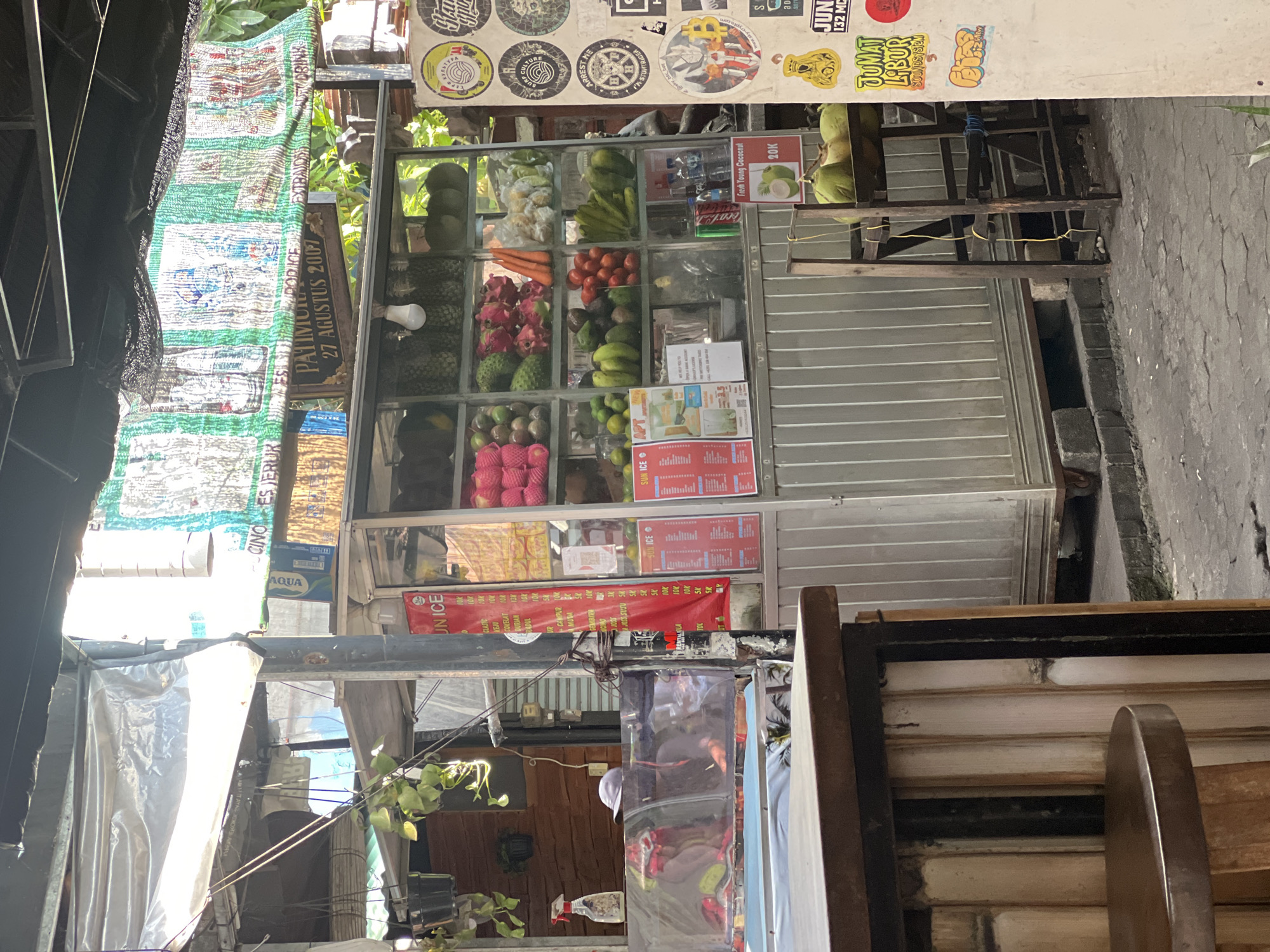 This image depicts an outdoor fruit stall in Kuta, Indonesia. The stall is constructed from metal and glass, displaying a variety of fresh fruits such as dragon fruit, pineapple, bananas, and avocados. There is also a sign that reads "Fresh Young Coconut" with a price of "20K". Several coconuts are visible on a wooden stand next to the stall. The stall is covered by a tarp and surrounded by other market elements, including a wooden counter in the foreground and various advertisements and stickers on the wall behind it. The ground is paved with cobblestones. The overall scene is a typical street-side market in a tropical urban environment.