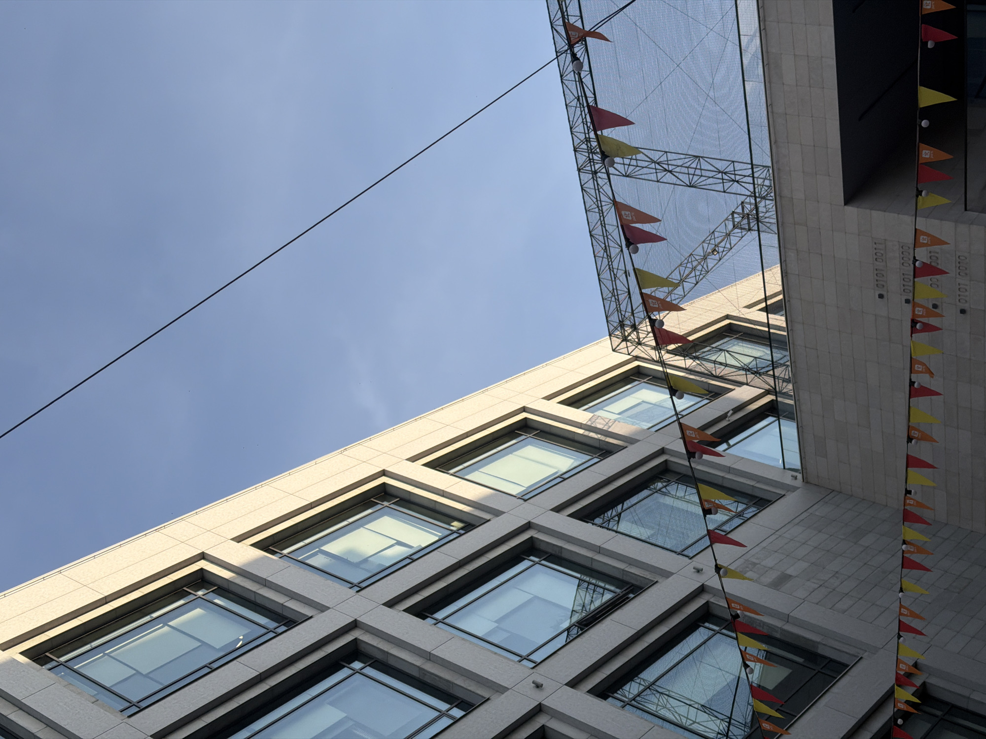 An upward-angled, architectural photograph captures two buildings against a clear blue sky. A utility line runs diagonally across the frame.
The building on the left features a pattern of diamond-shaped windows set into a light-colored, textured facade. The windows reflect the sky and other parts of the city.
The building on the right is darker in tone and appears to be made of a similar textured material. Strung between the buildings is a net-like structure adorned with small, colorful pennant flags. Additionally, a vertical line of the same flags is attached to the facade of the building on the right. The flags are red and yellow.
The location is likely in the city of Kazan, Russia. The scene is devoid of people. The lighting suggests it is daytime.