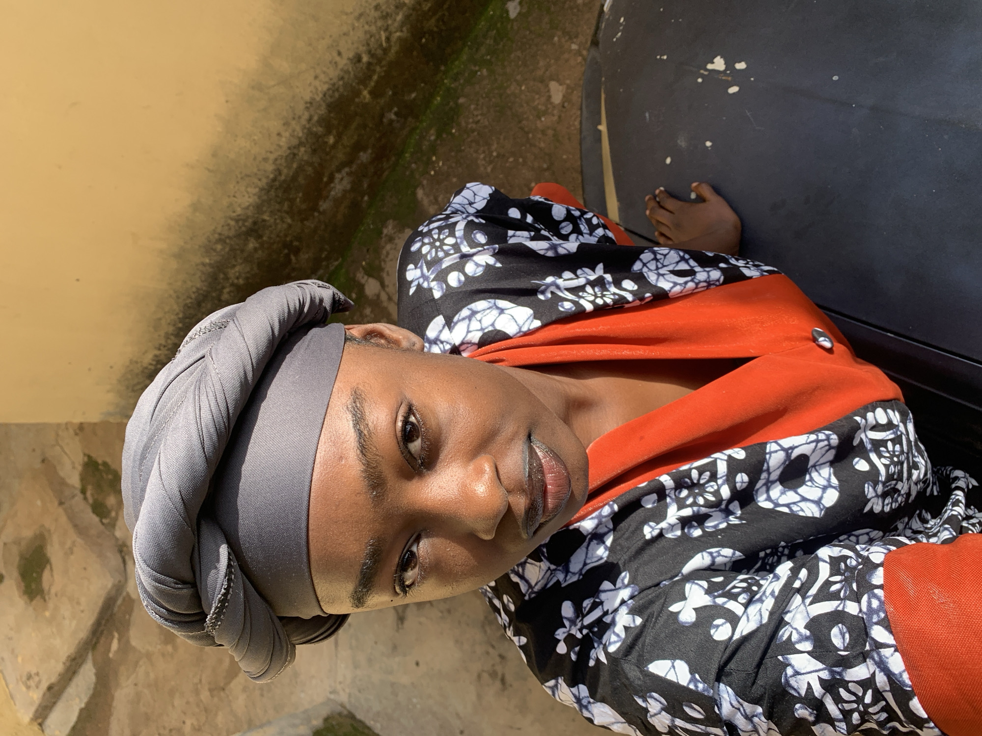 A woman is pictured from a high angle, looking upwards. She is wearing a grey headwrap and an orange and black patterned top. Her skin is dark, and she has dark eyes and lips. She is resting her hand on a dark, flat surface, possibly a car hood, which has white speckles on it. The background is a textured yellow wall and a grey concrete area with some green moss. The scene is outdoors. The woman appears to be in Rafin Par, Nigeria.