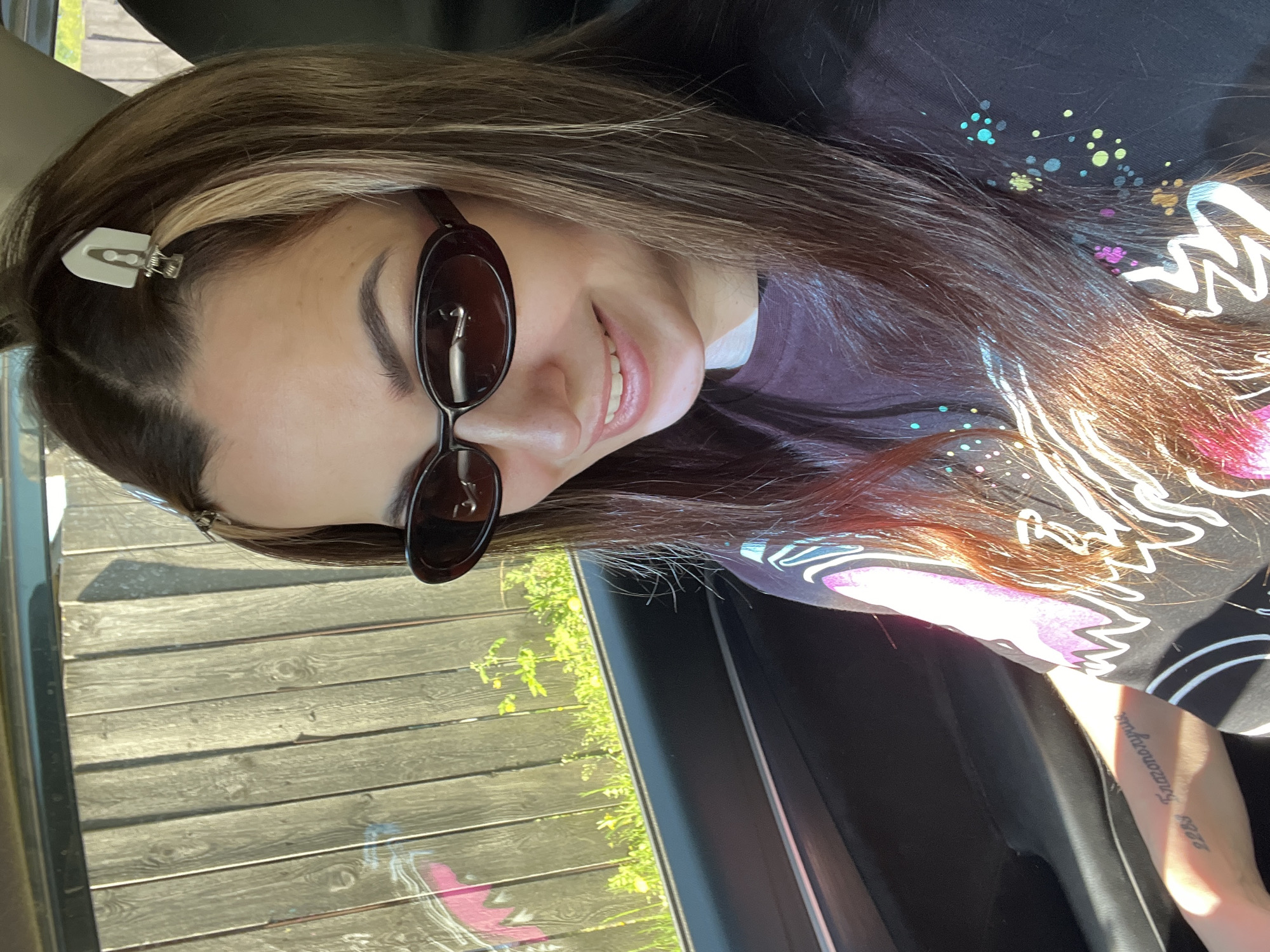 A woman with brown hair and sunglasses smiles while sitting in a car. Her hair has a clip in it, and she is wearing a black t-shirt with a colorful design. A tattoo is visible on her arm. The car window is open, revealing a wooden fence and some greenery outside. The location appears to be outdoors with ample sunlight. The location in the city is Unknown, Unknown.