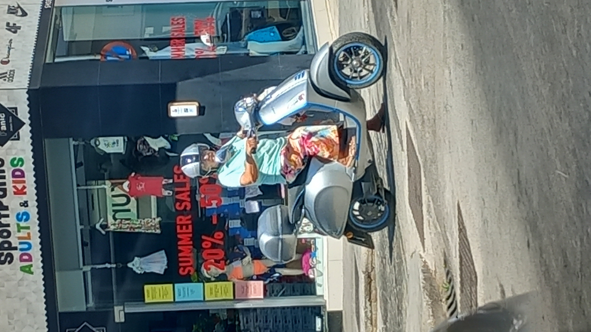 A person wearing a light blue polo shirt, a light grey helmet with black stripes, and brightly colored floral patterned pants is riding a silver scooter. The scooter has a silver storage box on the back and blue trim around the wheels. The person is looking forward as they ride. In the background, there is a storefront with a sign that reads "SportPanic ADULTS & KIDS" and "SUMMER SALES 20% - 50%". The storefront displays mannequins in swimwear. The scene appears to be on a street in Kos, Greece, based on the store's name and the overall aesthetic.