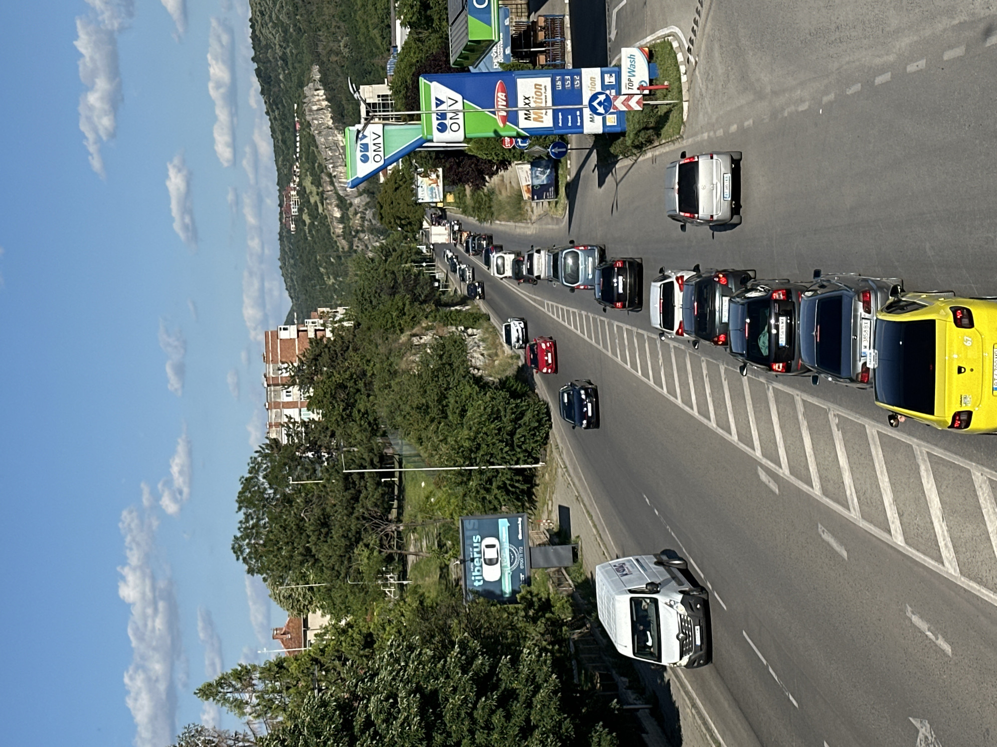 The image depicts a traffic scene on a multi-lane road in Veliko Tarnovo, Bulgaria. The road appears to be a main thoroughfare, with vehicles moving in a single direction. On the right side of the road, there is an OMV gas station with prominent signage. The surrounding environment includes trees, buildings, and a hilly landscape in the background. The sky is blue with scattered clouds.
Several vehicles are visible, including a white van on the left lane, followed by a queue of cars in the right lane. These cars vary in color and model. Some of the cars are dark colored, while others are white, silver, and red. A yellow car is also visible at the front of the queue.
In the distance, more vehicles can be seen, suggesting a moderate amount of traffic. The OMV gas station features fuel pumps and a car wash facility. A billboard advertising "tiberus s" is positioned on the left side of the road. The architecture of the buildings and the hilly terrain are characteristic of Veliko Tarnovo. The overall scene captures a typical urban traffic flow on a sunny day.