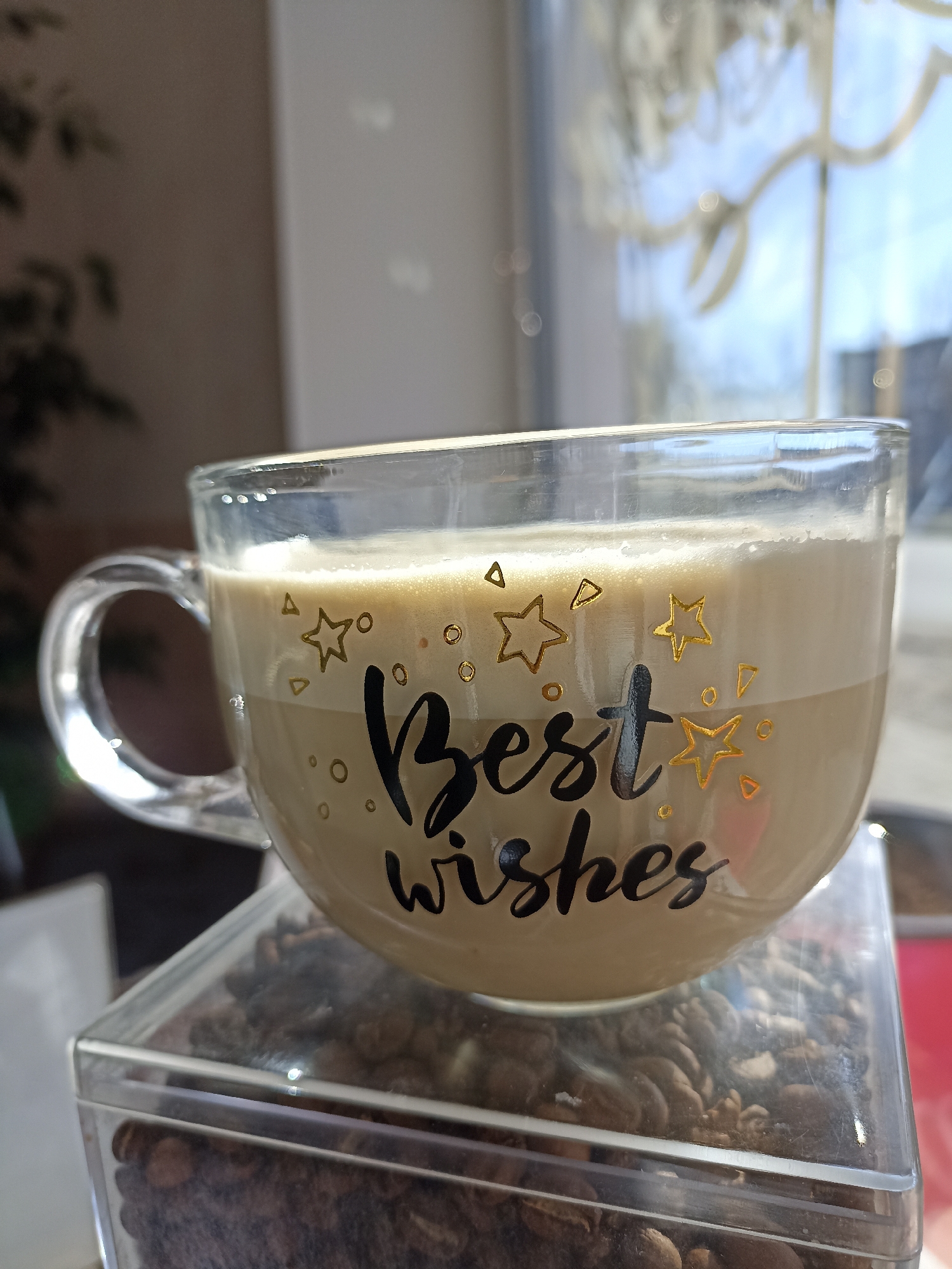 A close-up shot shows a clear glass mug filled with a latte, bearing the phrase "Best Wishes" and decorated with small golden stars and dots. The mug rests on a transparent plastic container filled with coffee beans. The background is blurred, but a window is visible with light filtering through, suggesting a daytime scene. The image appears to have been taken indoors. There is no information to specify the location of the image in the city of Chaikovsky, Russia.