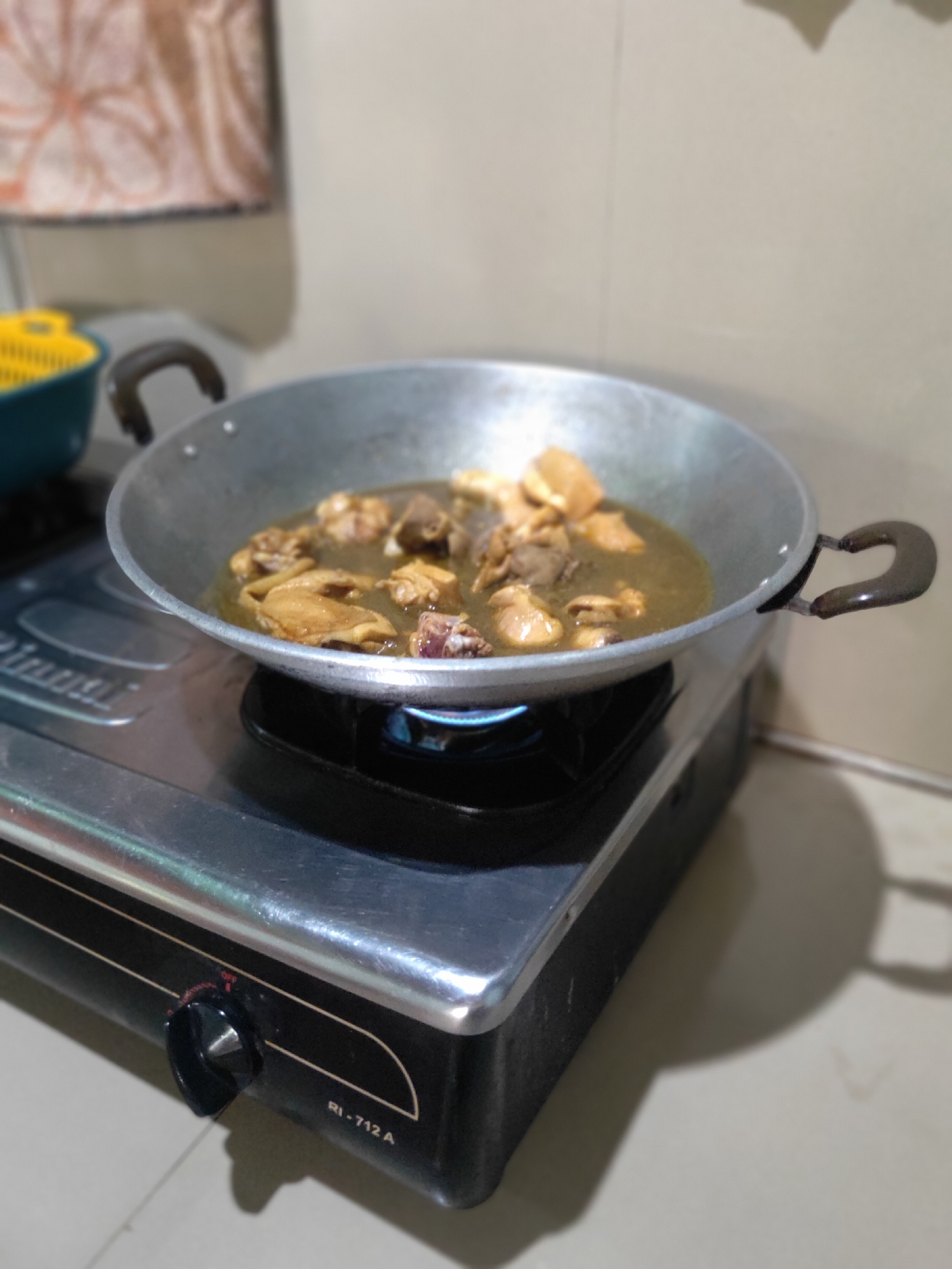 A close-up, eye-level shot captures a metal wok filled with what appears to be chicken pieces simmering in a dark broth. The wok rests on a black and silver gas stove, its burner emitting a blue flame. To the left of the wok, partially visible, is a teal bowl with a yellow colander. The background consists of a tiled wall. A knob on the stove, labeled "OFF," is visible in the lower portion of the frame. The overall scene suggests food preparation in a domestic kitchen setting.