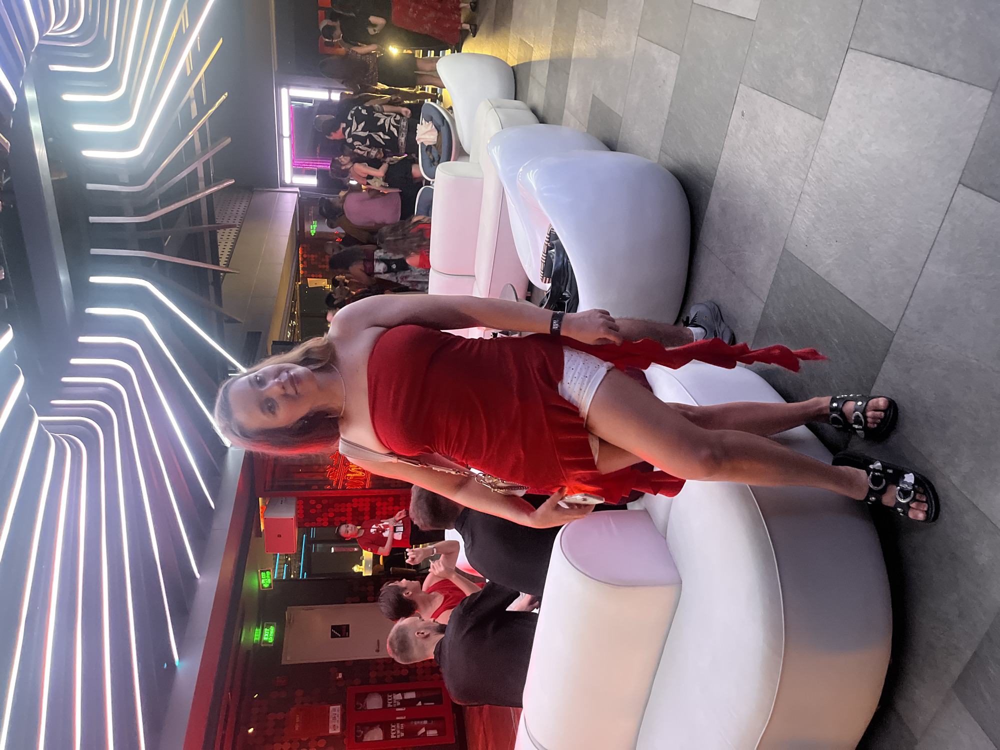 This media file captures a woman in a red strapless dress and white shorts, sitting on a white modular sofa in what appears to be an indoor entertainment venue. She is wearing black sandals with silver buckle details. The background shows a lively scene with multiple people, some standing and some seated, suggesting a social gathering or event. The venue is illuminated by bright, colorful LED lights that form an abstract pattern on the ceiling and walls, creating a modern and vibrant atmosphere. There are also signs with text in what appears to be Vietnamese, including one that says "EXIT" in green. The overall mood is festive and energetic. The lighting suggests it is evening or nighttime.