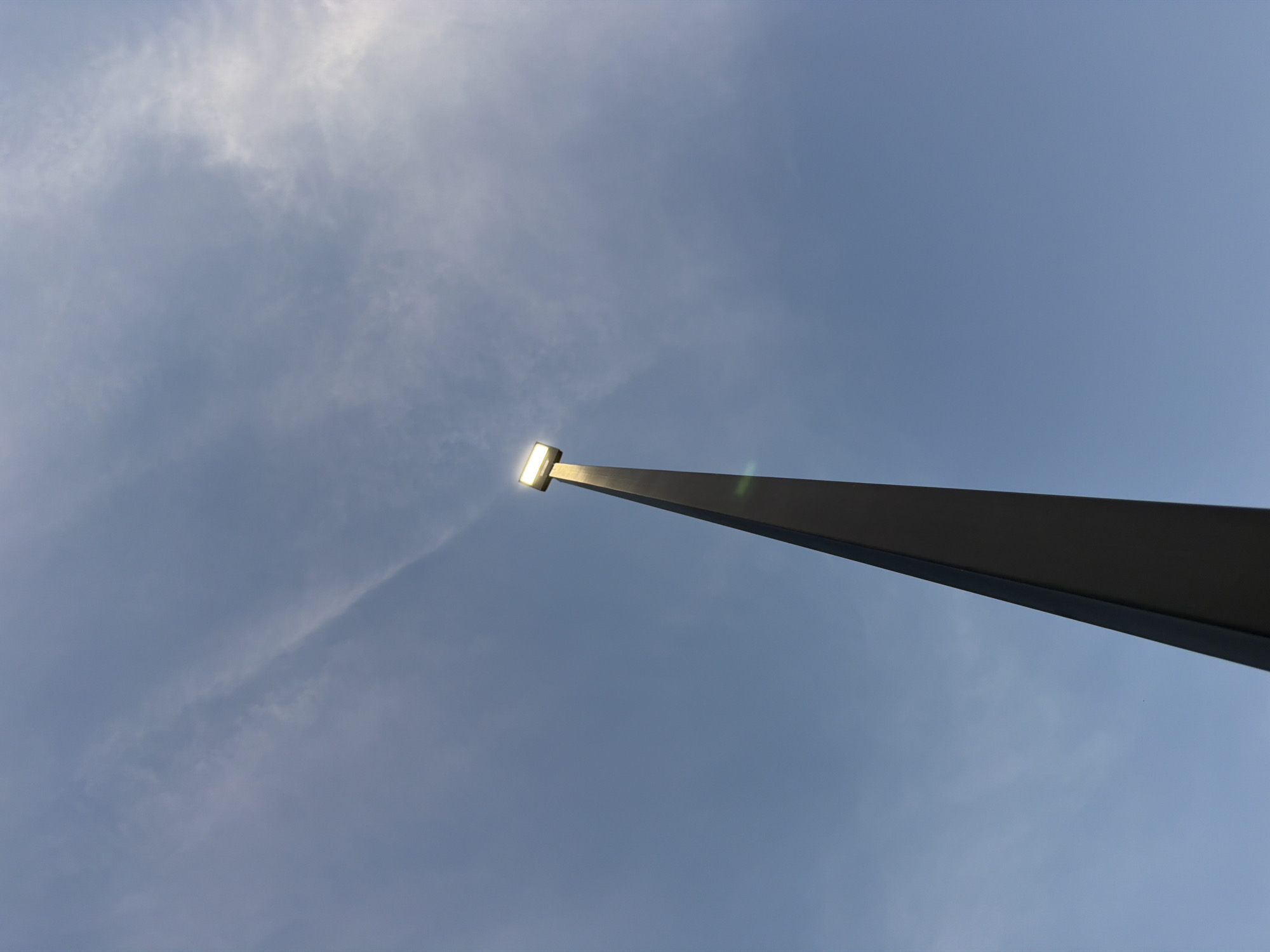 A street lamp stands tall against a pale blue sky with wispy clouds. The lamp is square and emits a bright white light, appearing to be on. The pole of the lamp is dark gray and tapers to a point as it extends upwards. There are no people or other objects visible in the frame. The image is taken from a low angle, looking up towards the sky. The location in the city of Cardwell, United States is unknown.